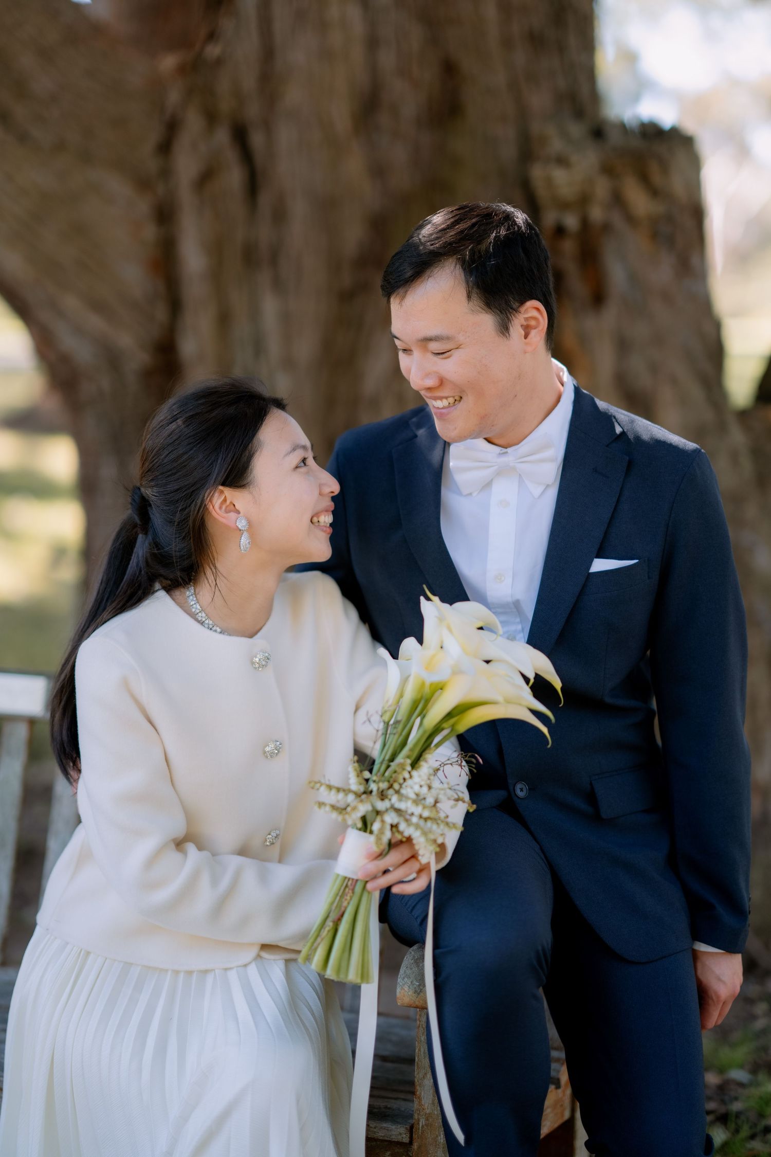 Bride in a cream jacket and pleated skirt laughs with her groom on a bench - candid Sydney elopement photography