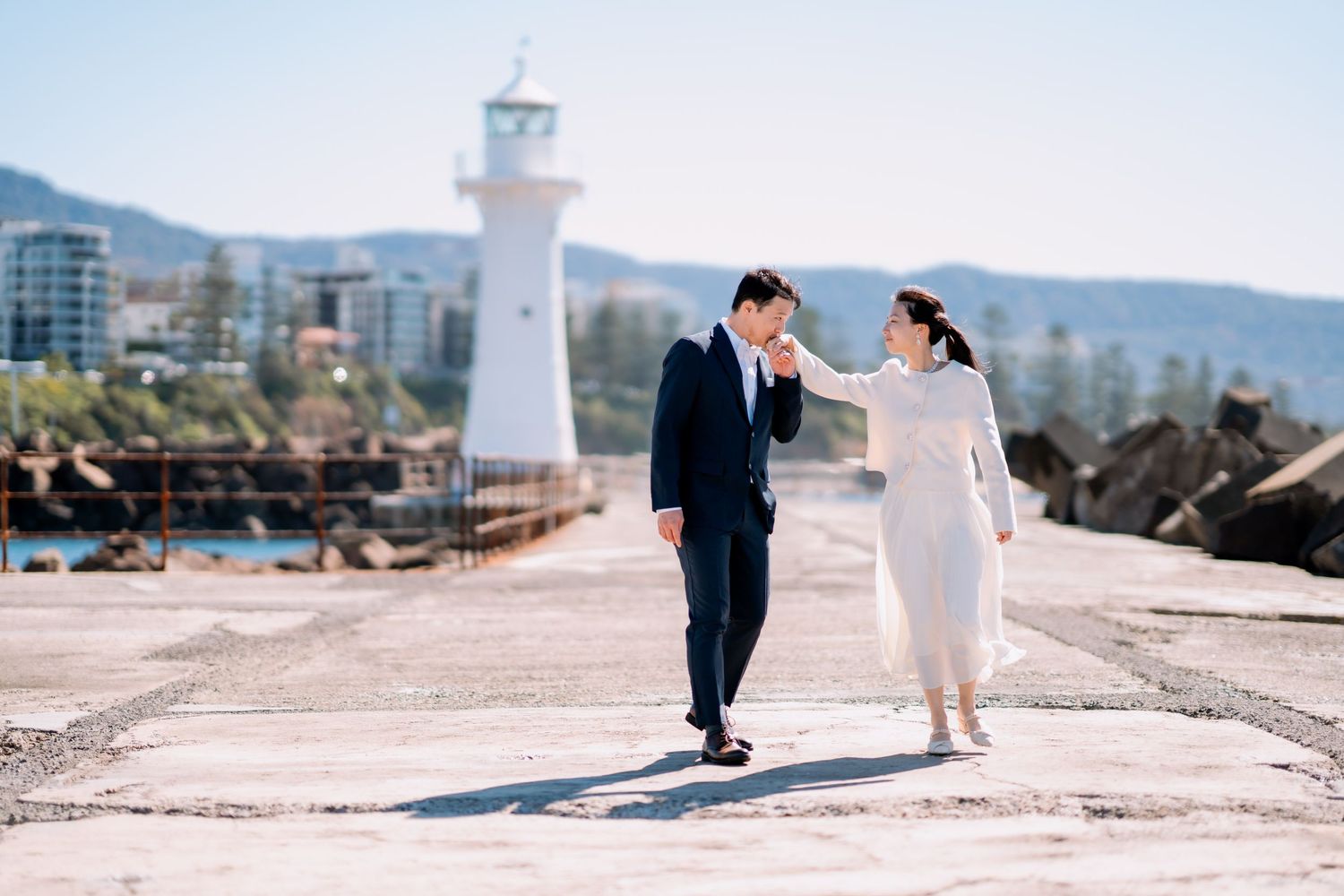 Groom holds his bride's hand on a stone pier beside a white lighthouse - coastal elopement photography