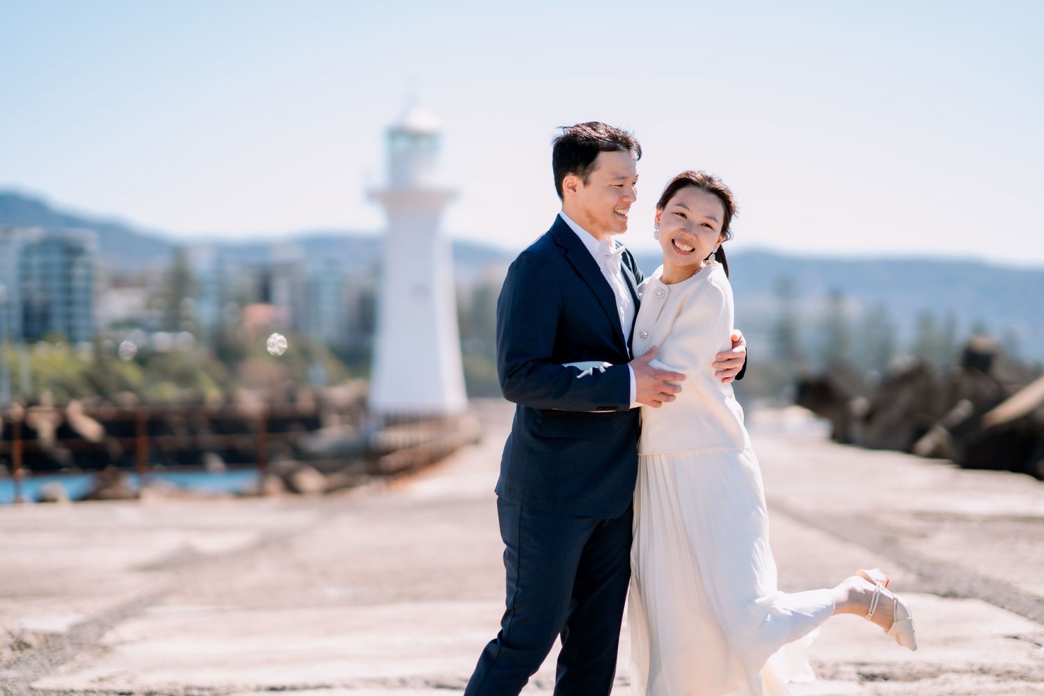 Couple embrace on a sunny harbour breakwater with a white lighthouse and hills - seaside elopement portraits