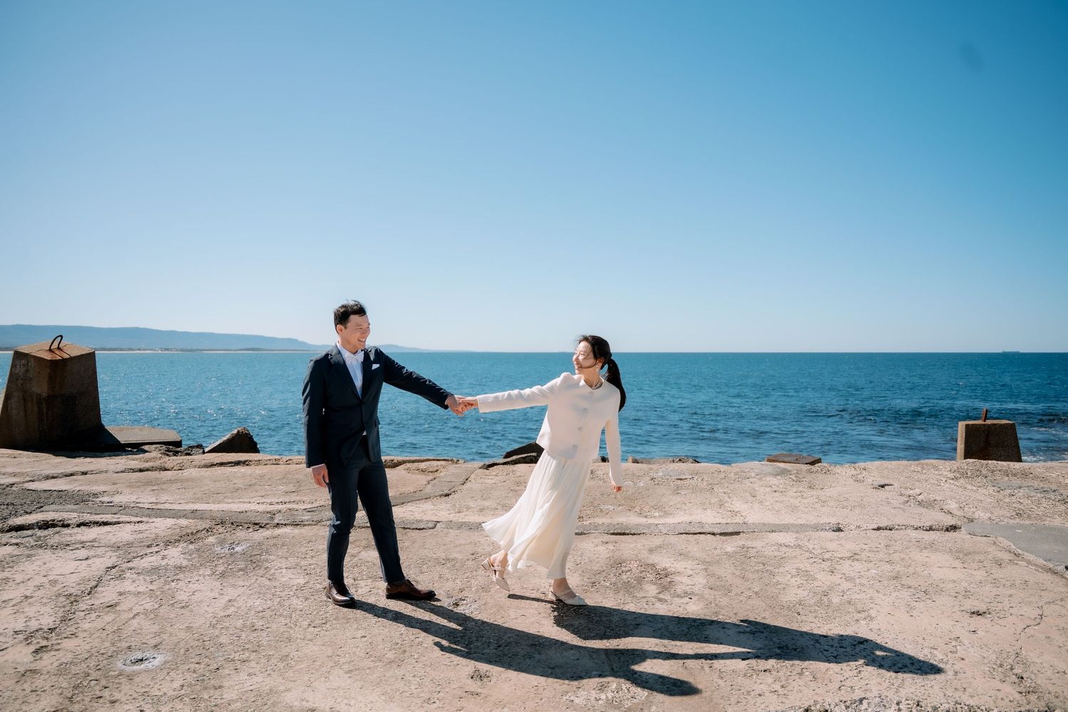 Bride and groom walk hand in hand along a sunny rocky seaside ledge - coastal elopement photographer