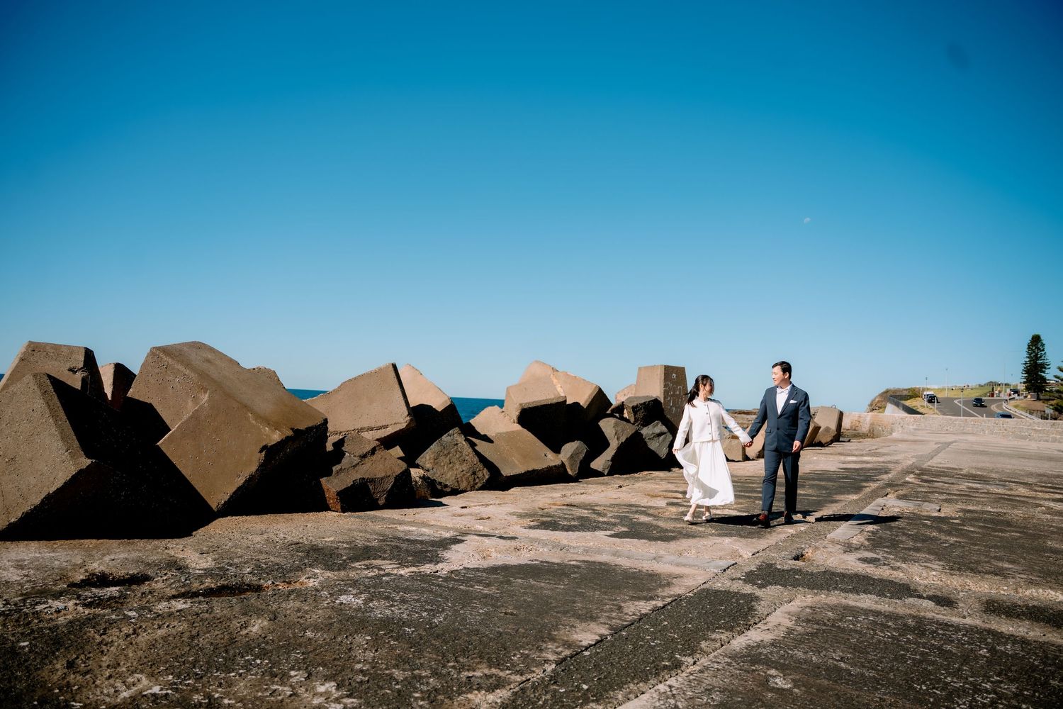 Newlyweds walk across a rocky seaside pier beside large stones - ocean elopement photography, NSW