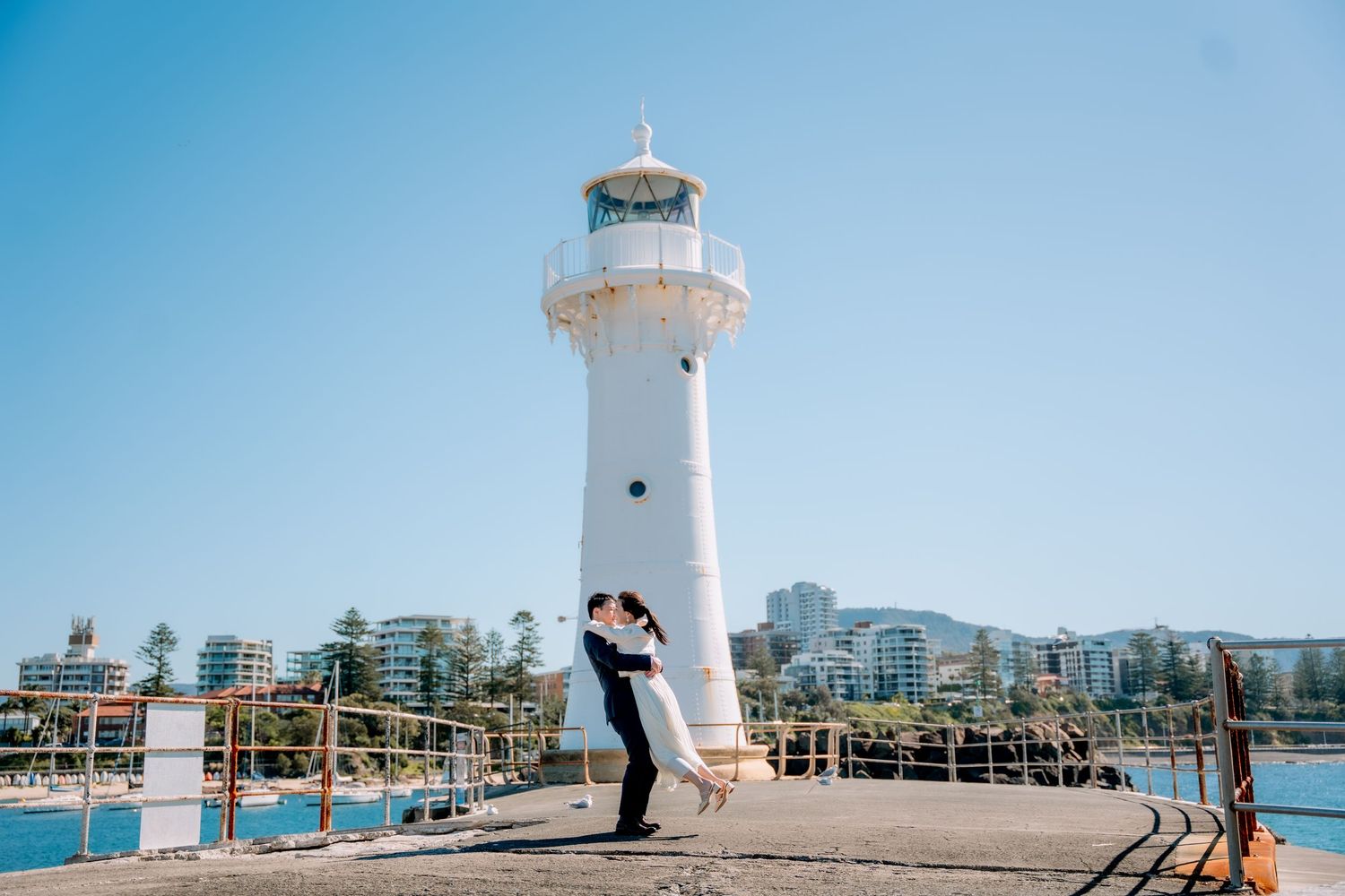 Bride and groom kiss on a stone pier beside a white lighthouse - romantic seaside elopement photography