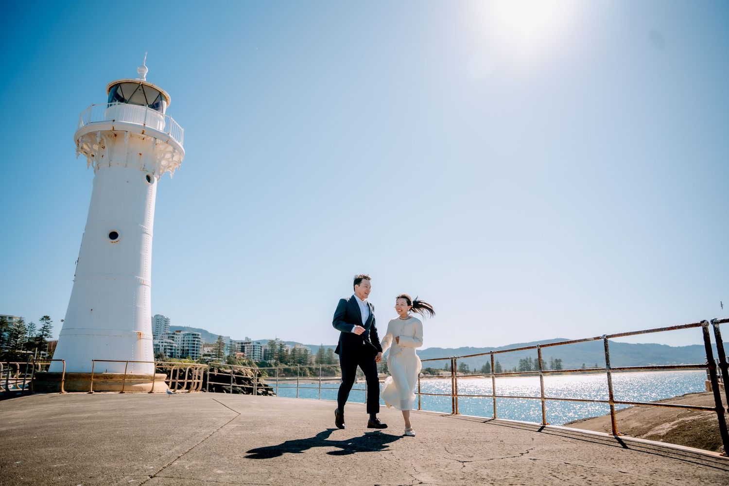 Couple walk hand in hand along a pier past a white lighthouse under a bright sky - NSW coastal elopement