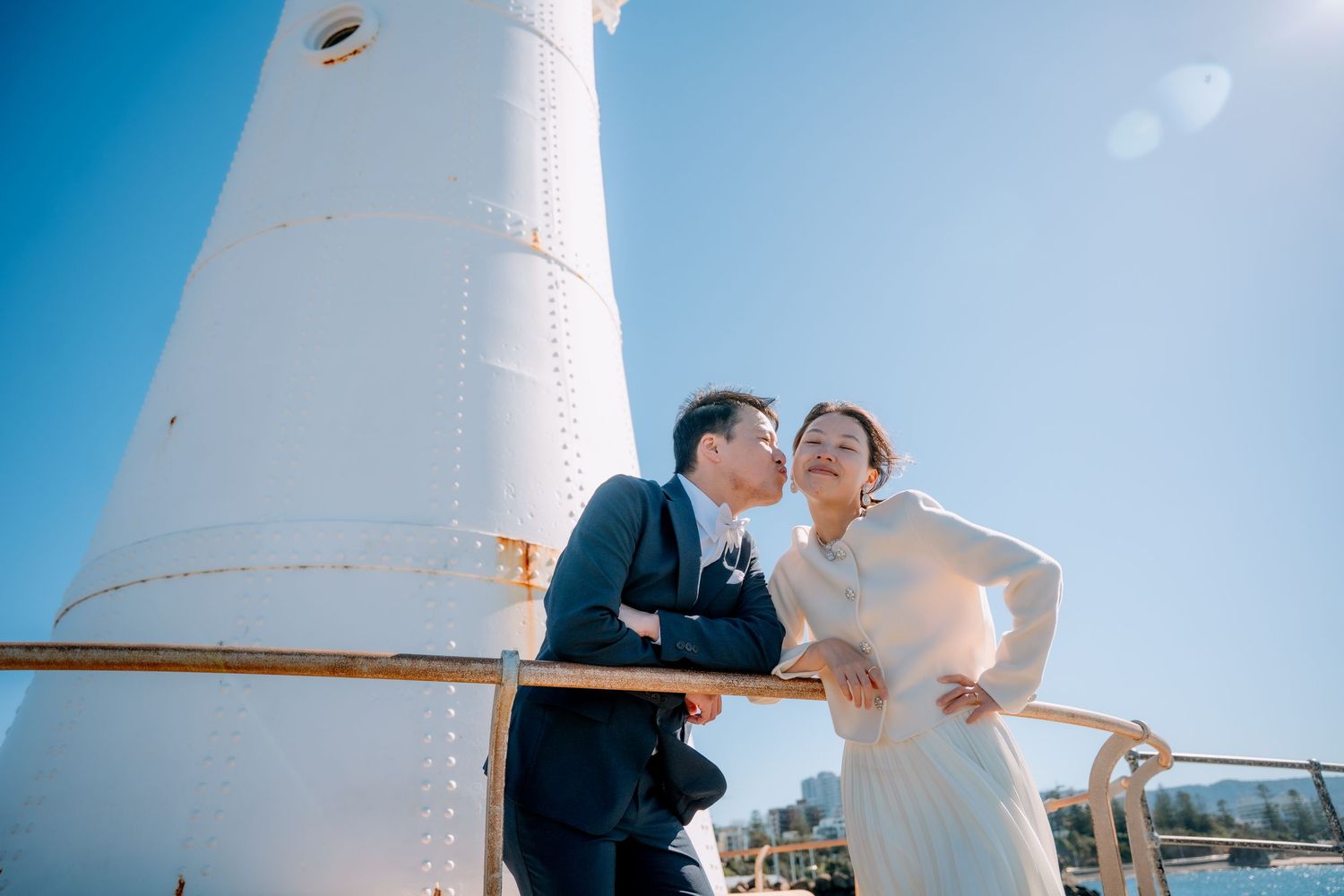 Groom kisses his bride's cheek at a railing beside a white lighthouse - elopement couple portraits by the sea