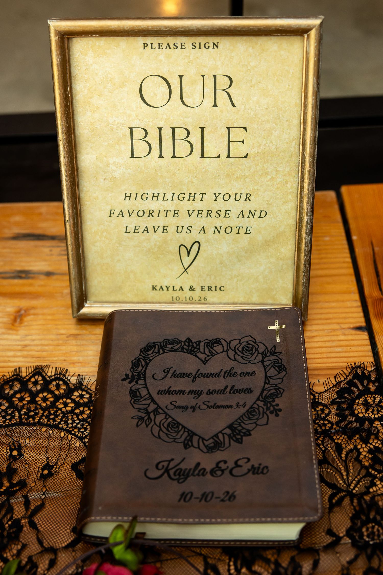 Wedding guest book sign reading Our Bible beside a engraved leather Bible for Kayla and Eric, set on a wooden table with black lace.