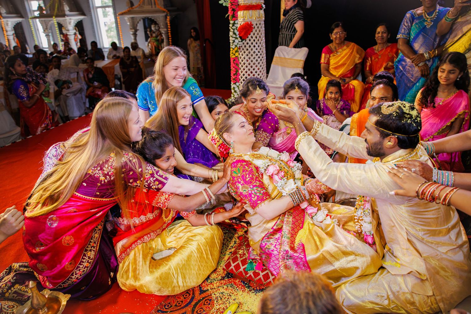Family members in colorful traditional Indian attire gather around a bride and groom on a red carpet during a joyful wedding ceremony.