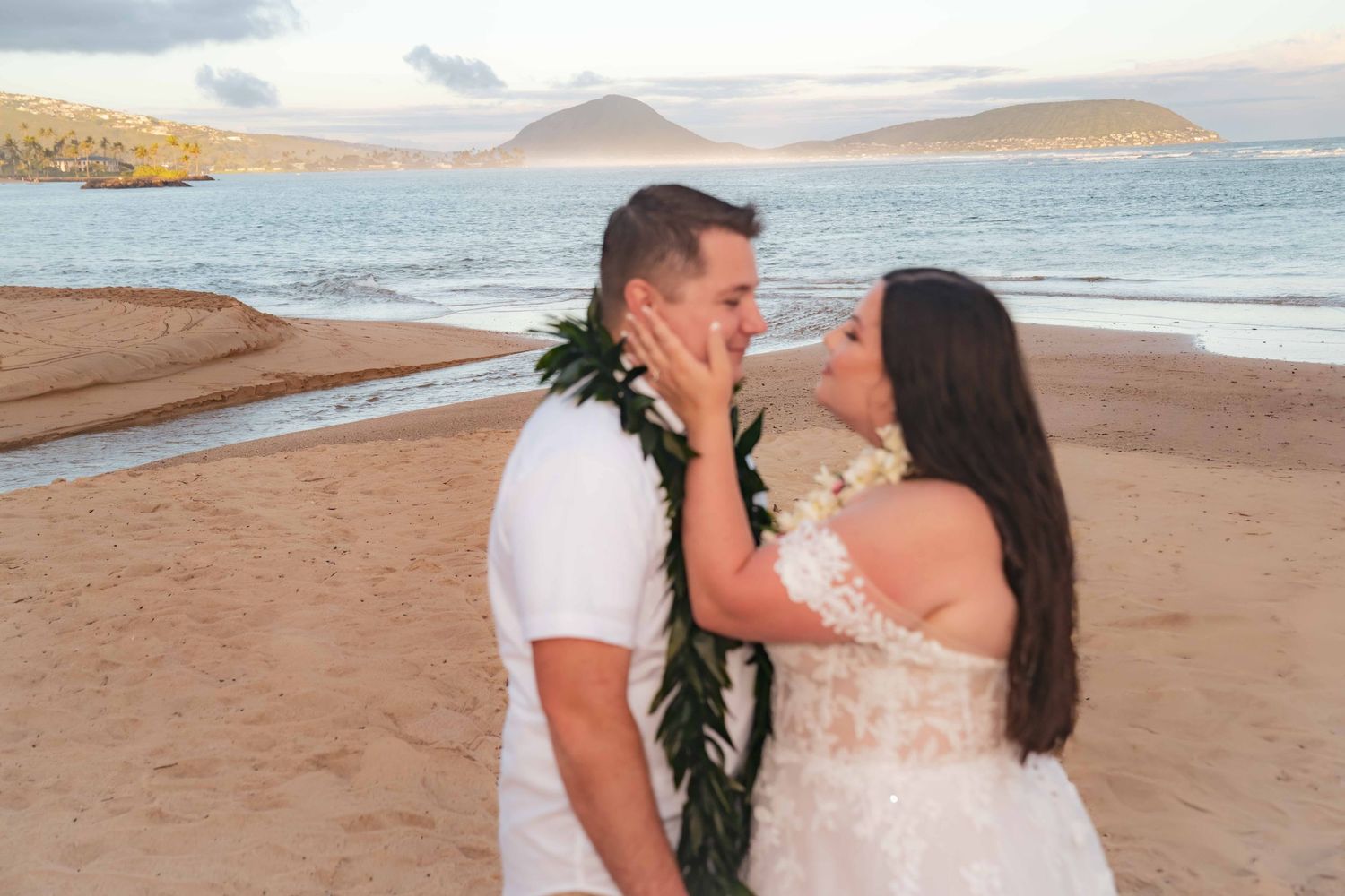 Bride in a lace wedding dress touches the face of a groom wearing a green leaf lei on a sandy beach with ocean and hills behind them.