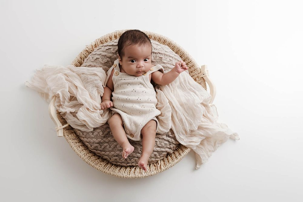 Newborn baby in cream knit romper lying in woven basket with soft beige fabric on white background.