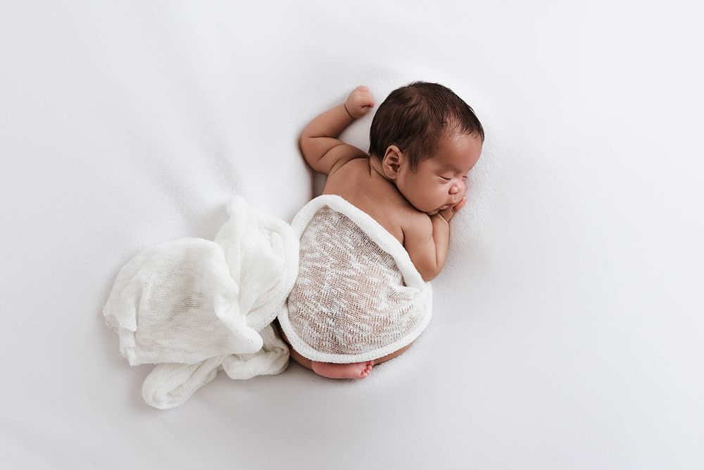 Newborn baby sleeping on white blanket with white lace wrap draped over back.