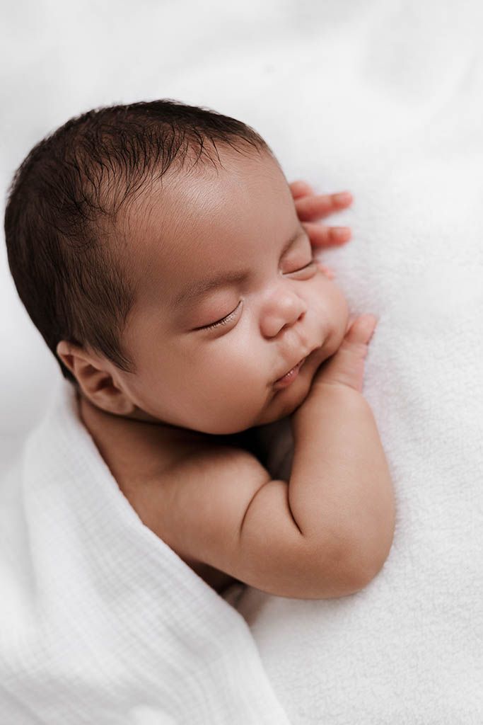 Close-up of sleeping newborn baby with dark hair resting on hands in white wrap.