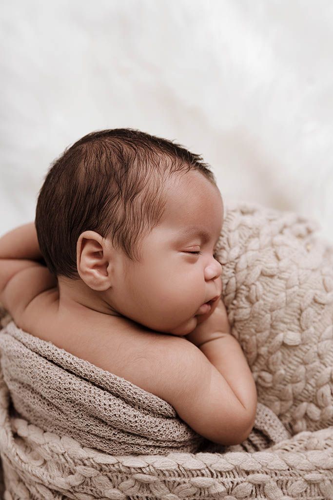 Peaceful newborn baby sleeping curled up in a knit basket wrapped in a soft beige blanket.