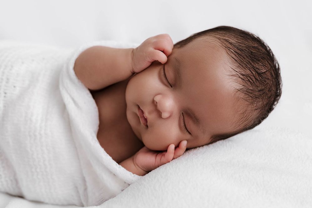 Peaceful newborn baby sleeping wrapped in a soft white swaddle blanket with tiny hands near face.