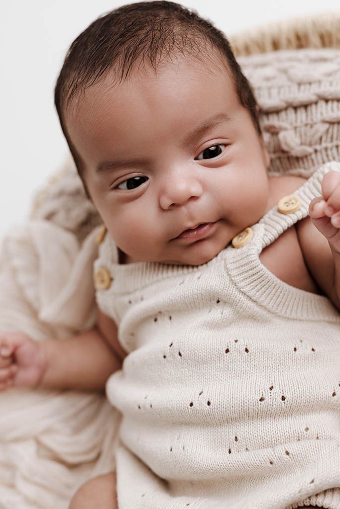 Adorable newborn baby in cream knit romper sitting in woven basket.