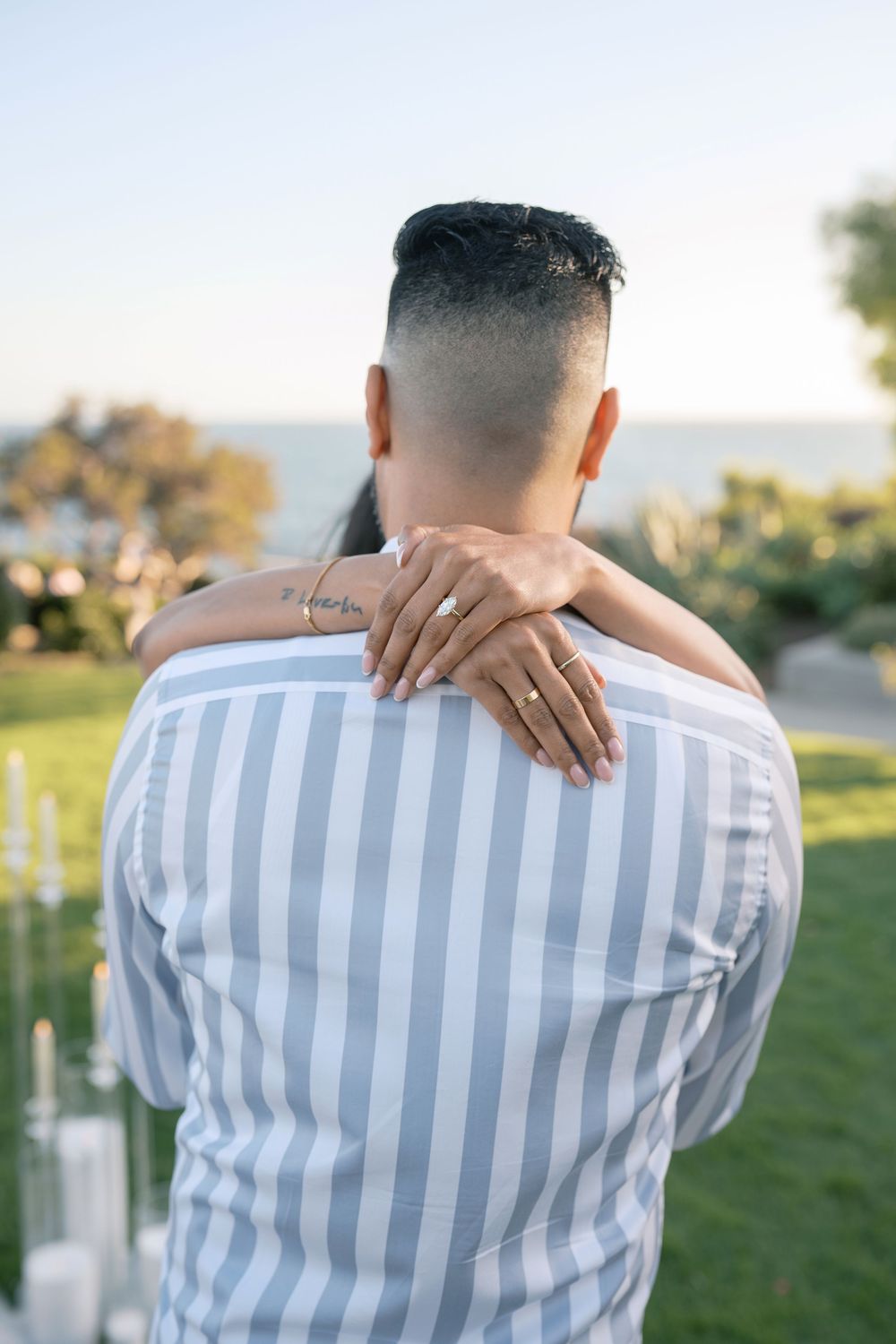 Couple embracing outdoors with engagement ring visible, ocean view backdrop, man in striped shirt.