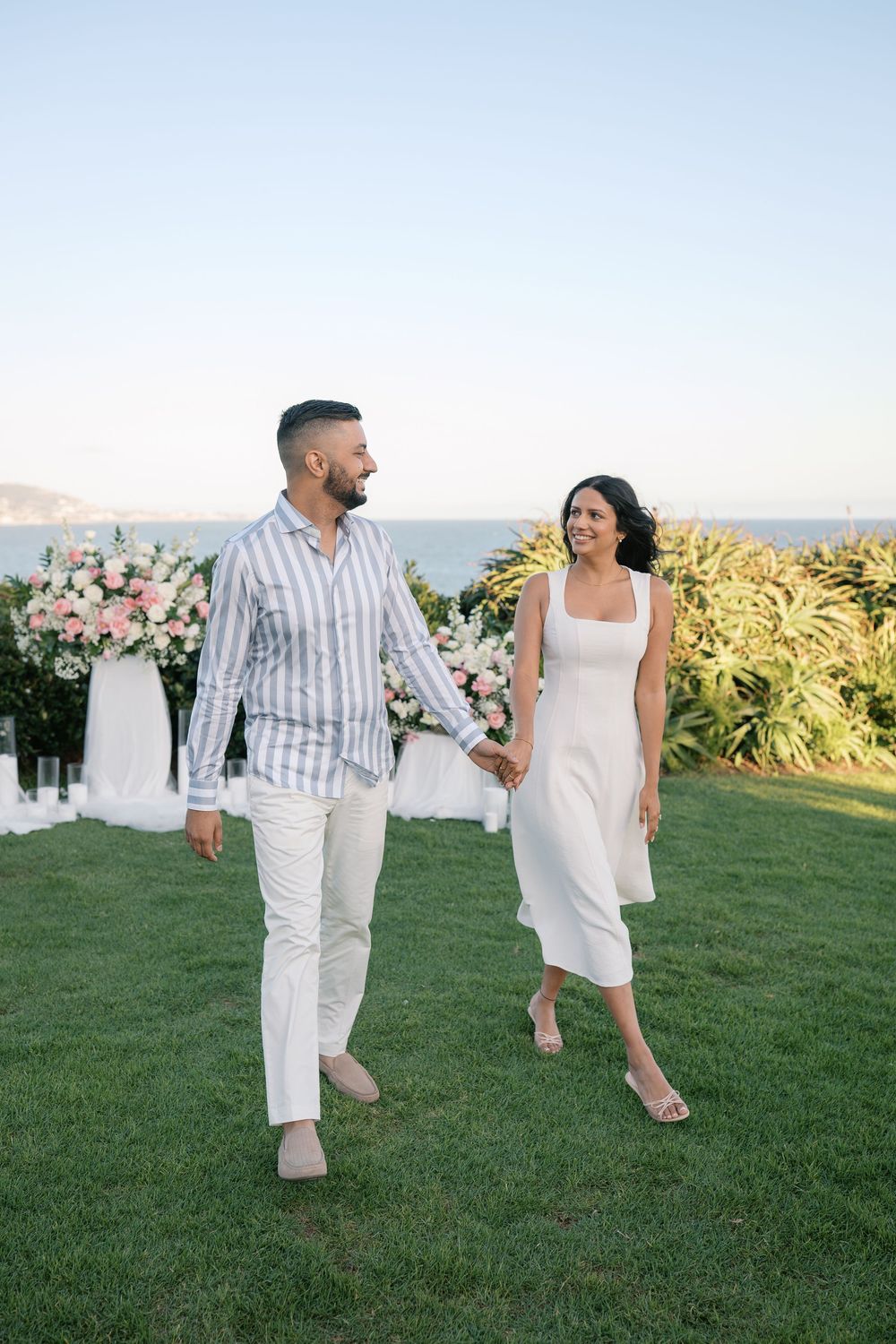 Couple in white outfits holding hands and smiling while walking on green lawn with ocean view and floral arrangements.