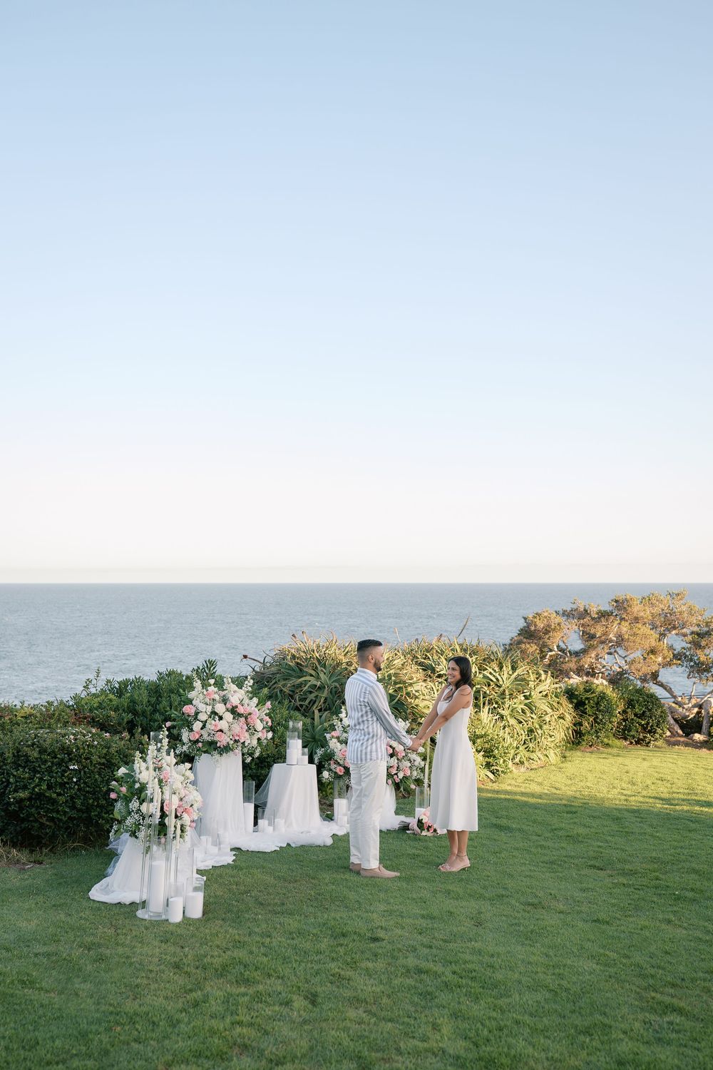 Couple holds hands during an intimate outdoor wedding ceremony on a cliffside lawn overlooking the ocean with white floral arrangements.