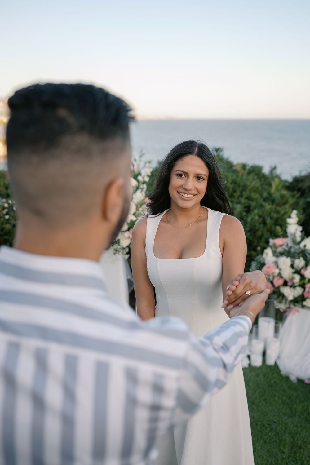 Smiling bride in white dress holds hands with groom during romantic outdoor proposal by the ocean at sunset.