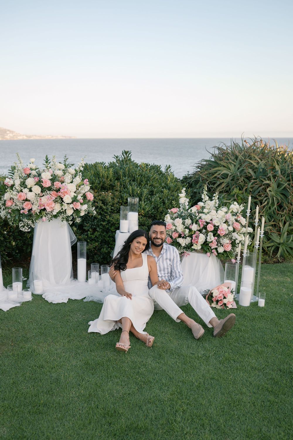 Couple sitting on grass surrounded by pink floral arrangements and candles with ocean view at sunset.