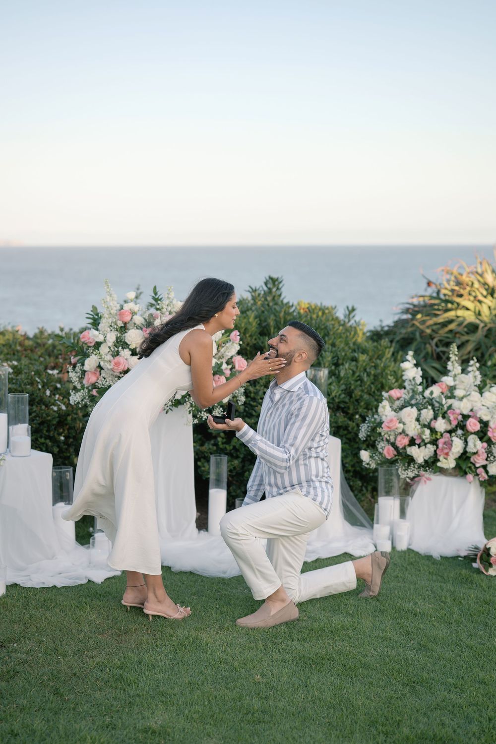 Man proposing on one knee to woman outdoors with pink flowers and ocean backdrop at dusk.