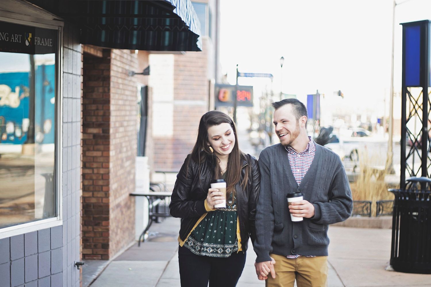 A smiling couple walks along a city sidewalk, each holding coffee cups on a sunny day.
