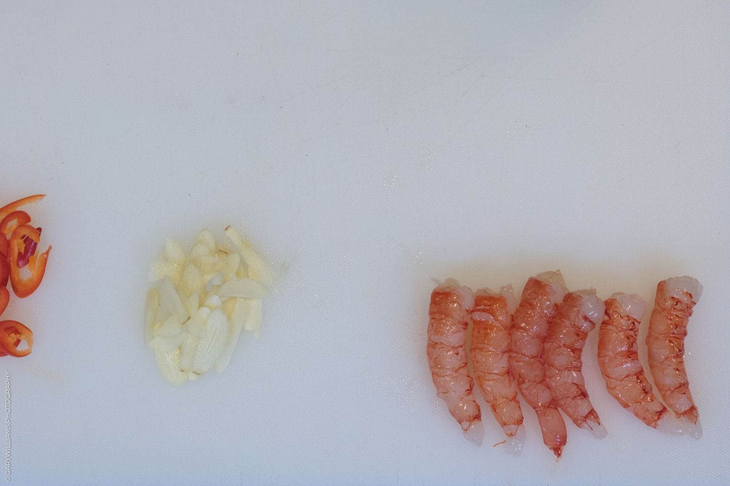Sliced red chili peppers, minced garlic, and peeled raw shrimp arranged in a row on a white cutting board.