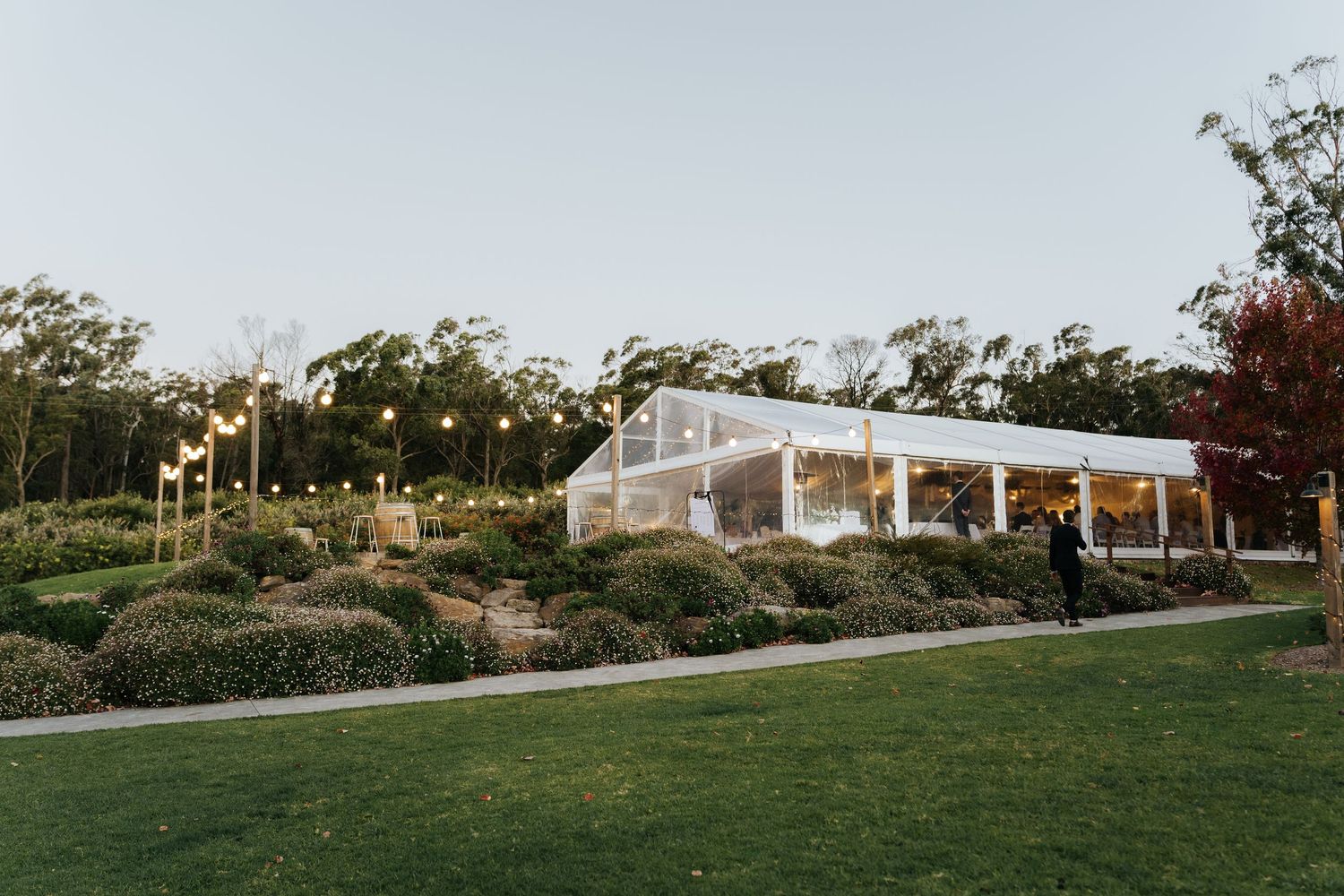 Elegant glass marquee wedding venue surrounded by manicured gardens and string lights at dusk.