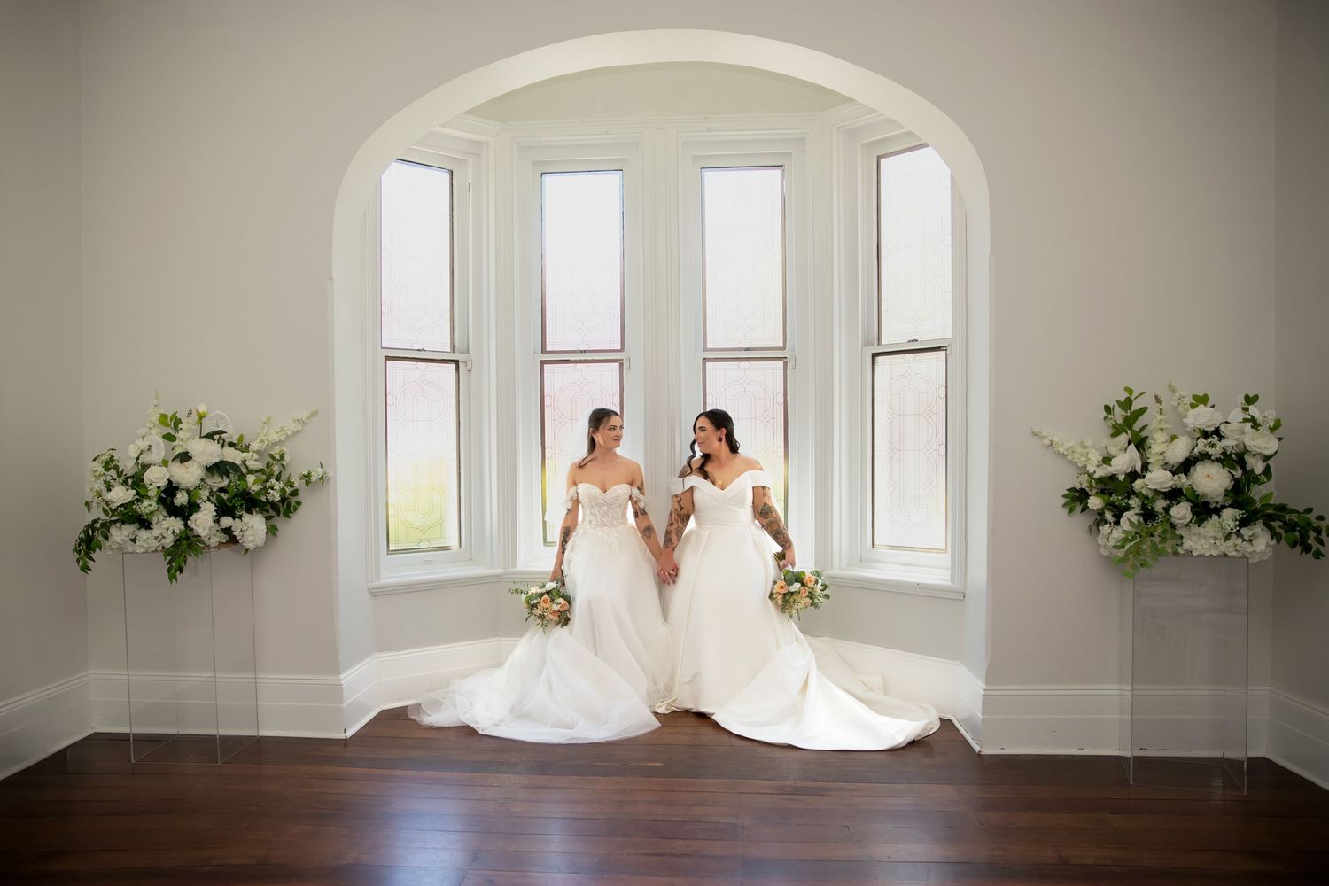 Two brides in white gowns hold hands by arched windows with floral arrangements at their wedding ceremony.