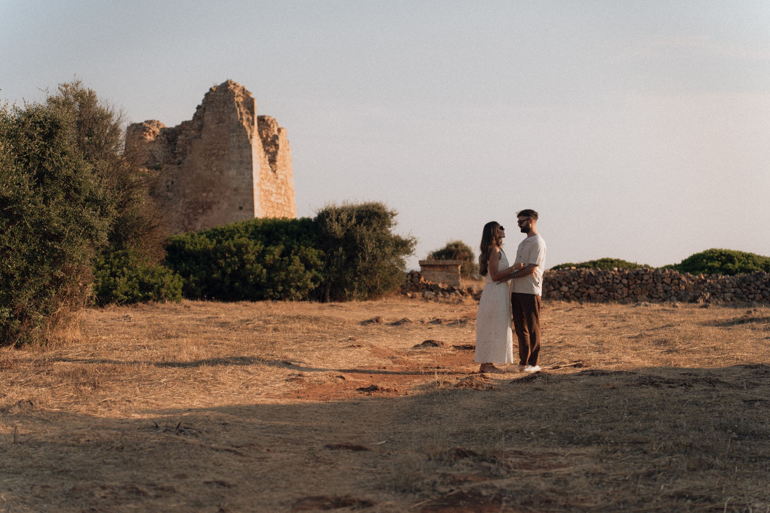 Romantic couple standing together near historic stone tower ruins in warm sunset light outdoors.