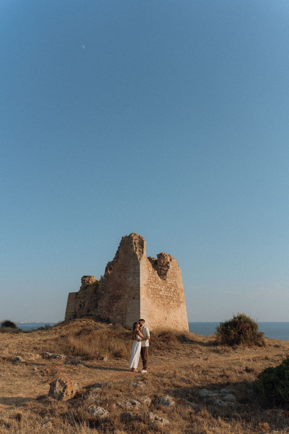 Couple embracing in front of ancient stone ruins on a hilltop overlooking the sea under a clear blue sky.