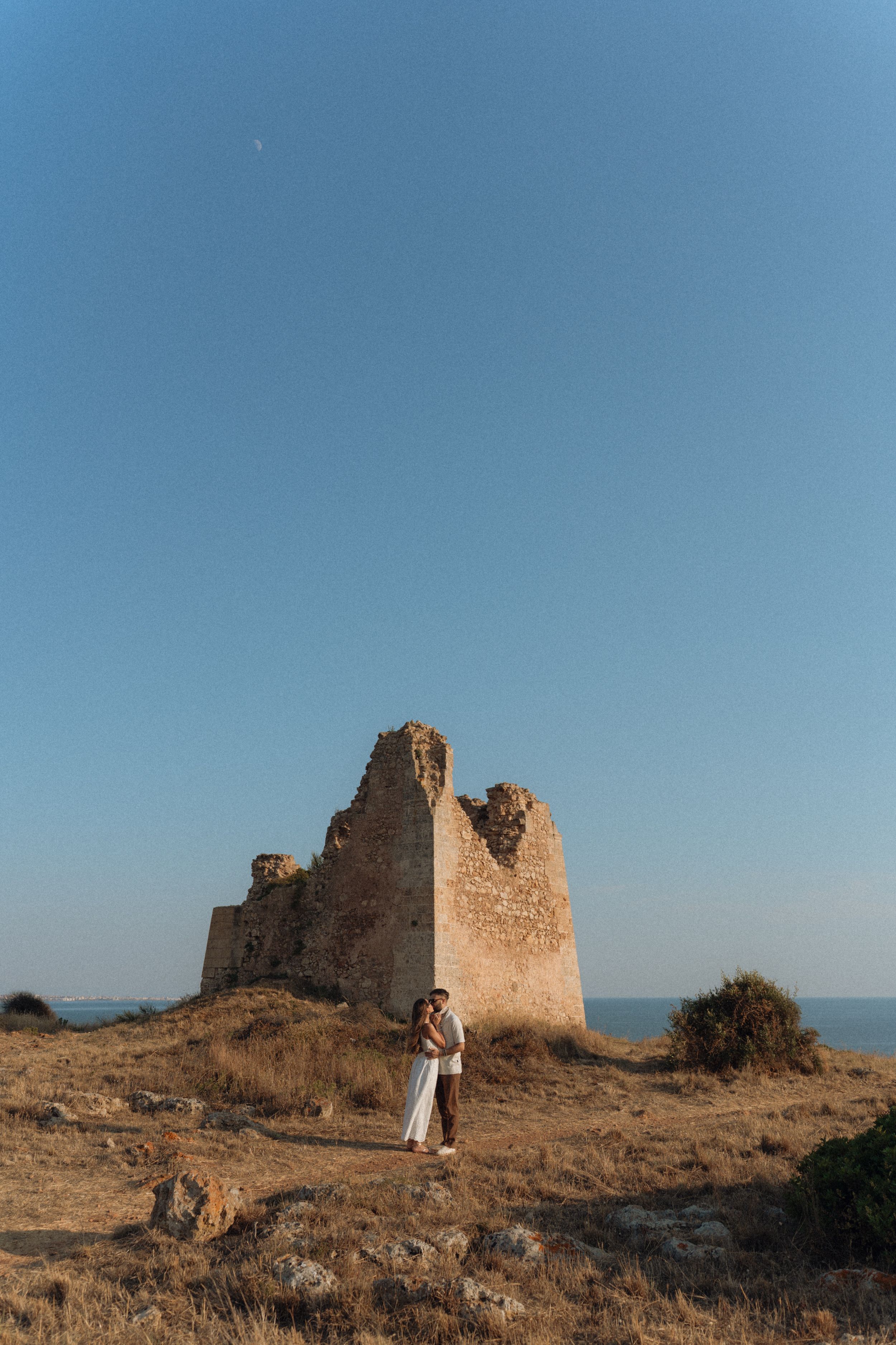 Couple embracing in front of ancient stone ruins on a hilltop overlooking the sea under a clear blue sky.
