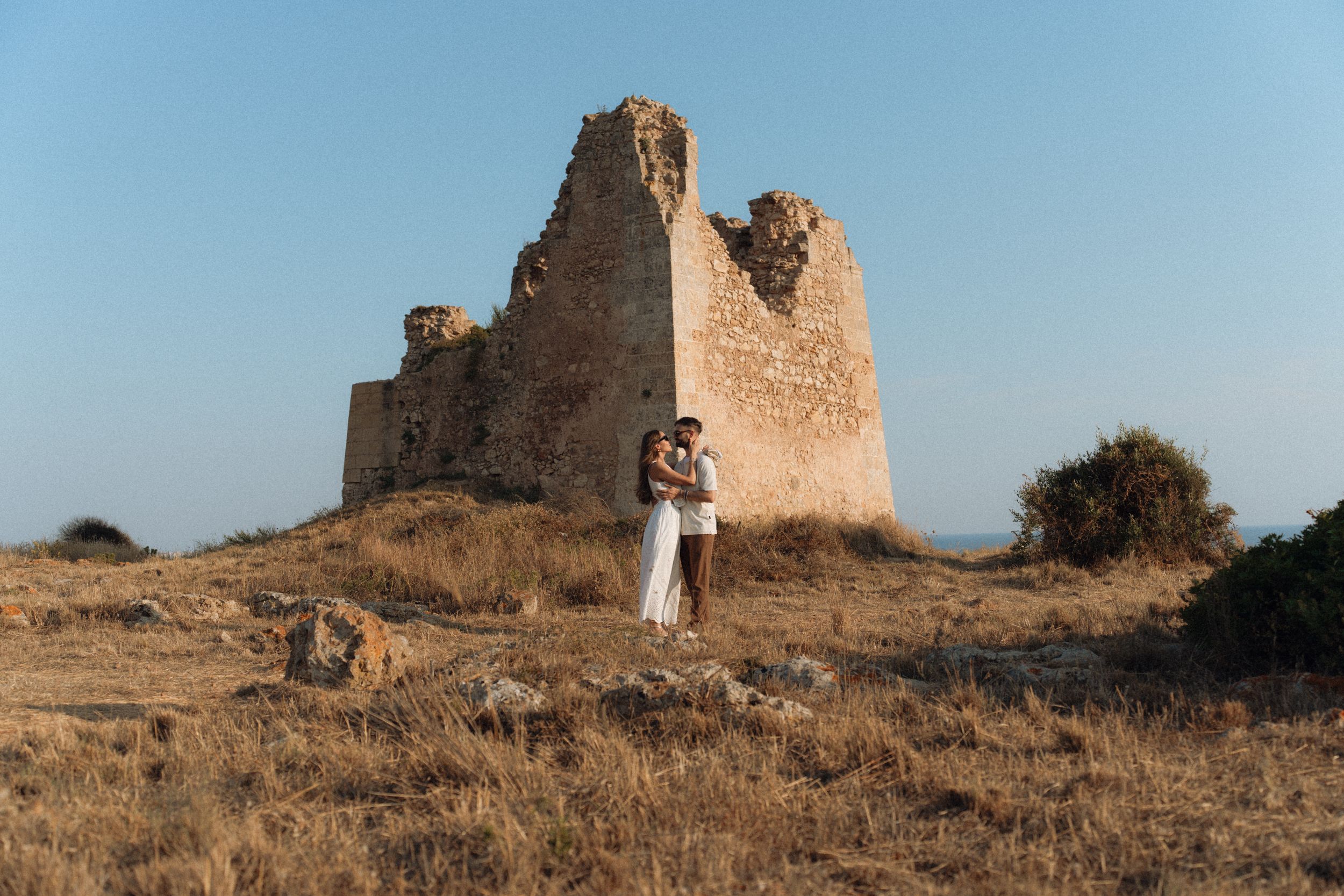 Couple embracing romantically in front of ancient stone ruins on a golden dry hillside under a clear blue sky.