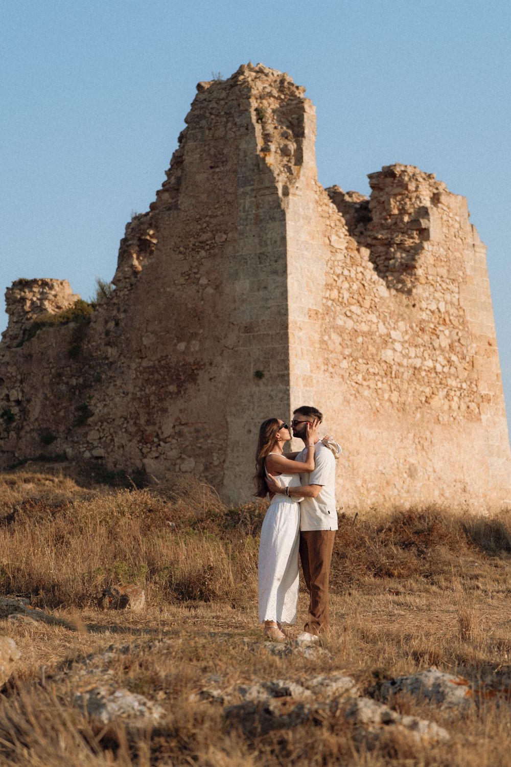 Romantic couple embracing in front of ancient stone ruins under a clear blue sky.