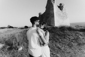 Black and white photo of a romantic couple kissing near historic stone tower ruins in an open field.