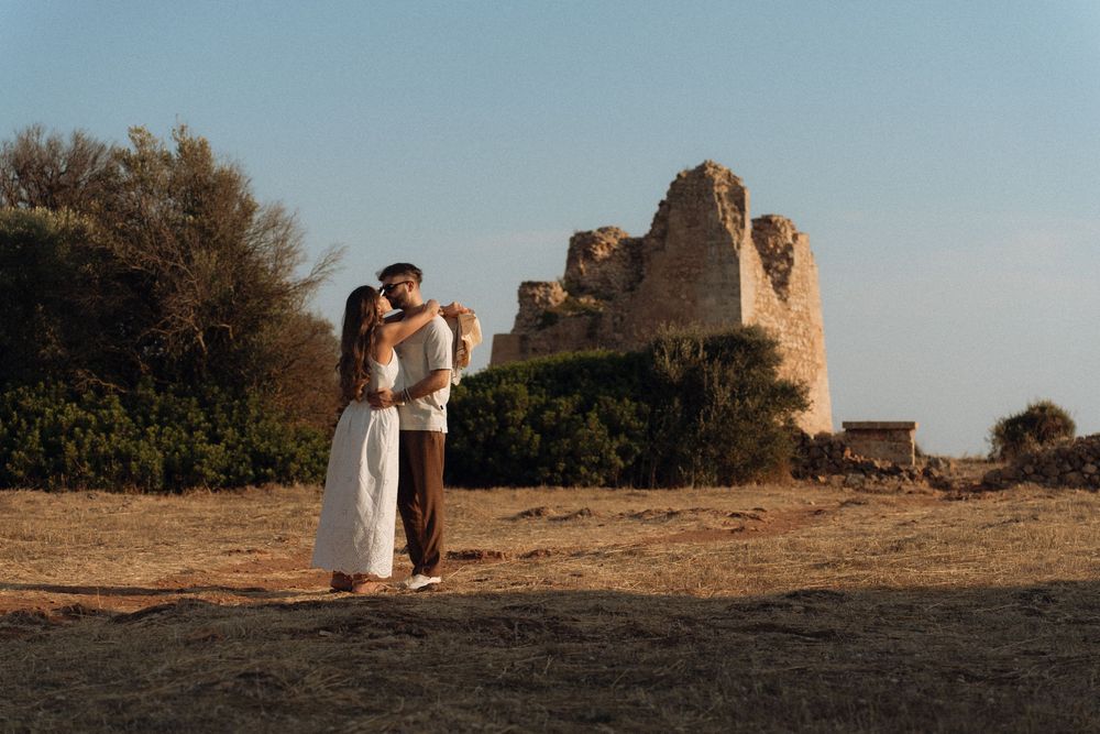 Couple embracing at golden hour near ancient stone ruins in an open field with trees and clear blue sky.