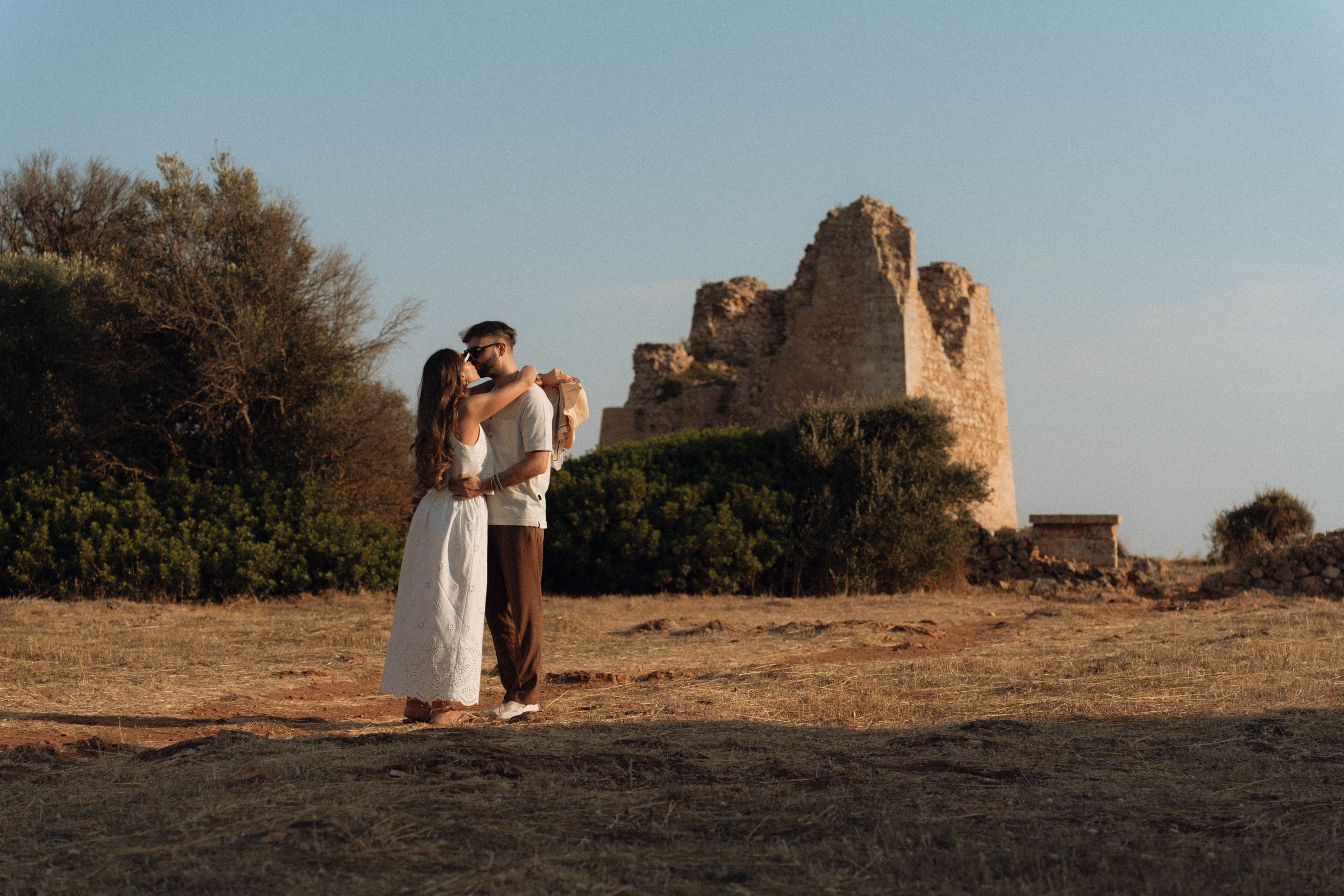 Couple embracing at golden hour near ancient stone ruins in an open field with trees and clear blue sky.
