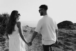 Black and white image of couple holding hands walking toward ocean horizon on grassy cliff.