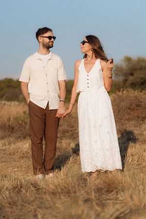 Couple holding hands in a dry field, both wearing sunglasses, woman in white dress, man in beige shirt.