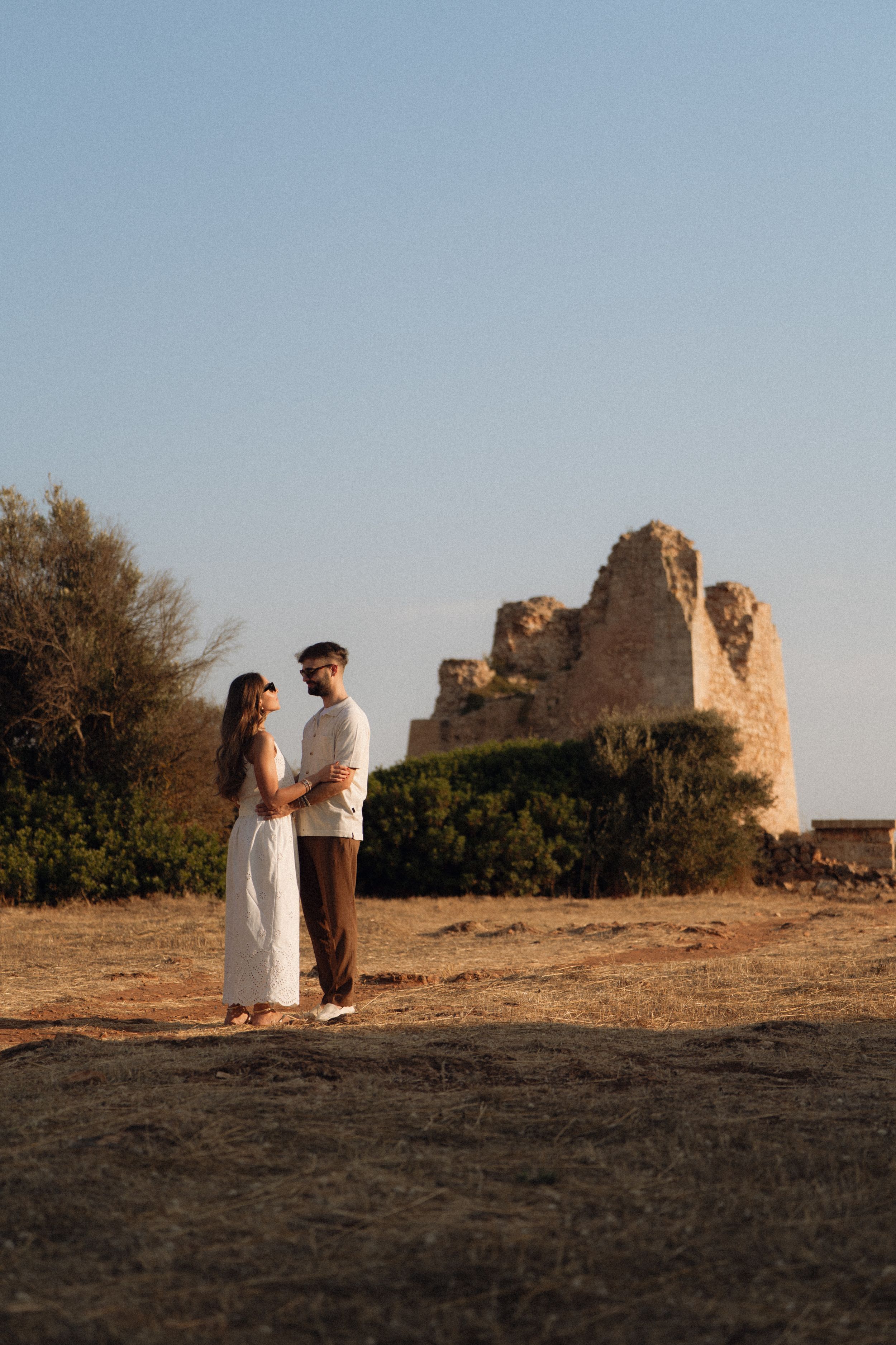 Couple holding hands in white dress and dark pants before ancient stone ruins at golden hour.