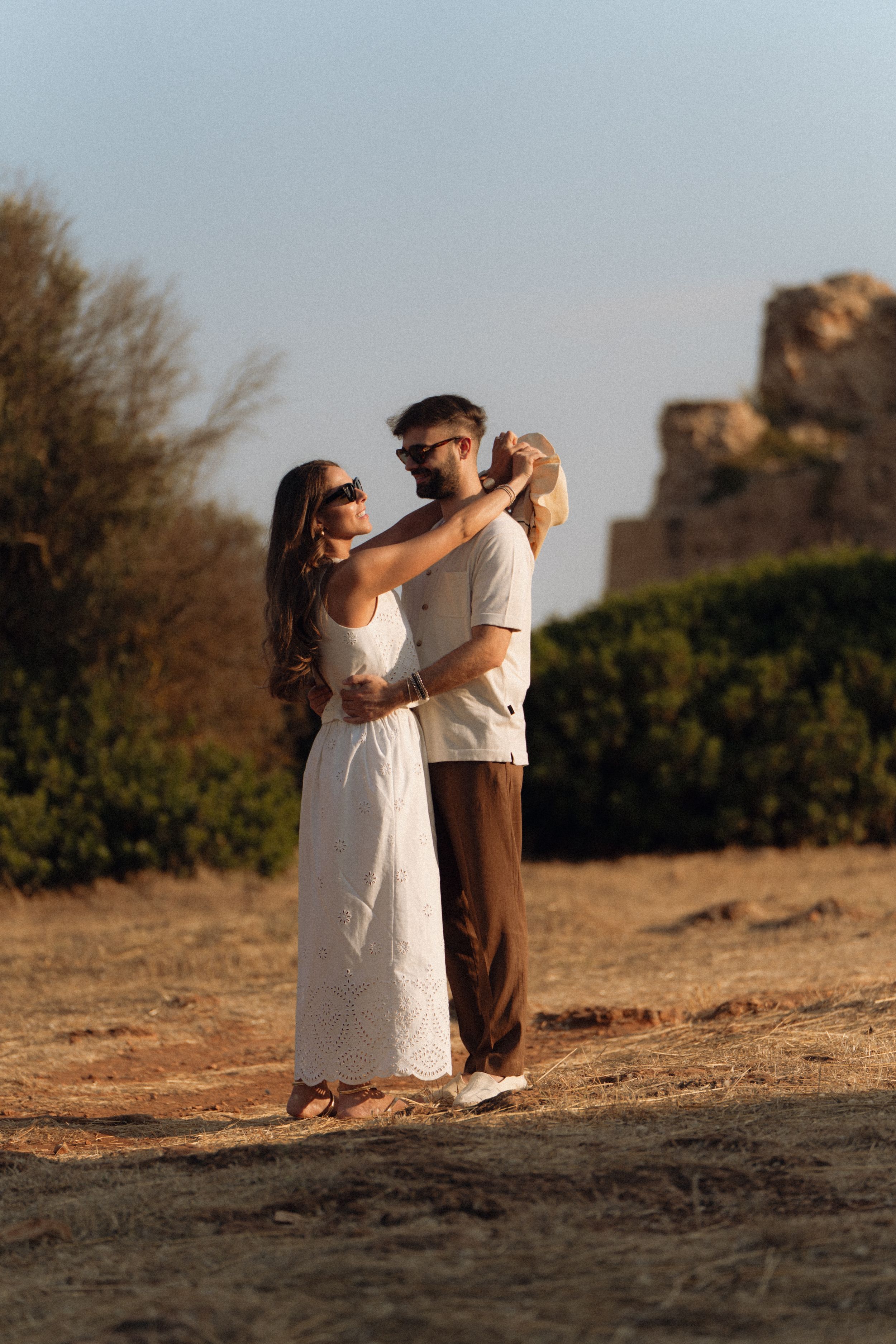 Romantic couple embracing outdoors at golden hour, woman in white dress, man in brown pants, ancient ruins in background.