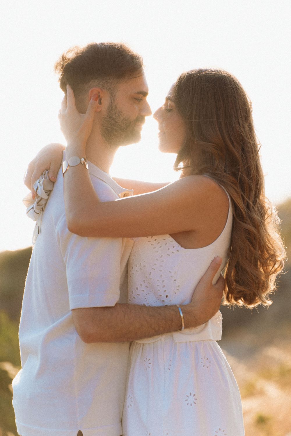 Young couple in white outfits sharing a tender moment, embracing face-to-face in warm golden sunlight outdoors.