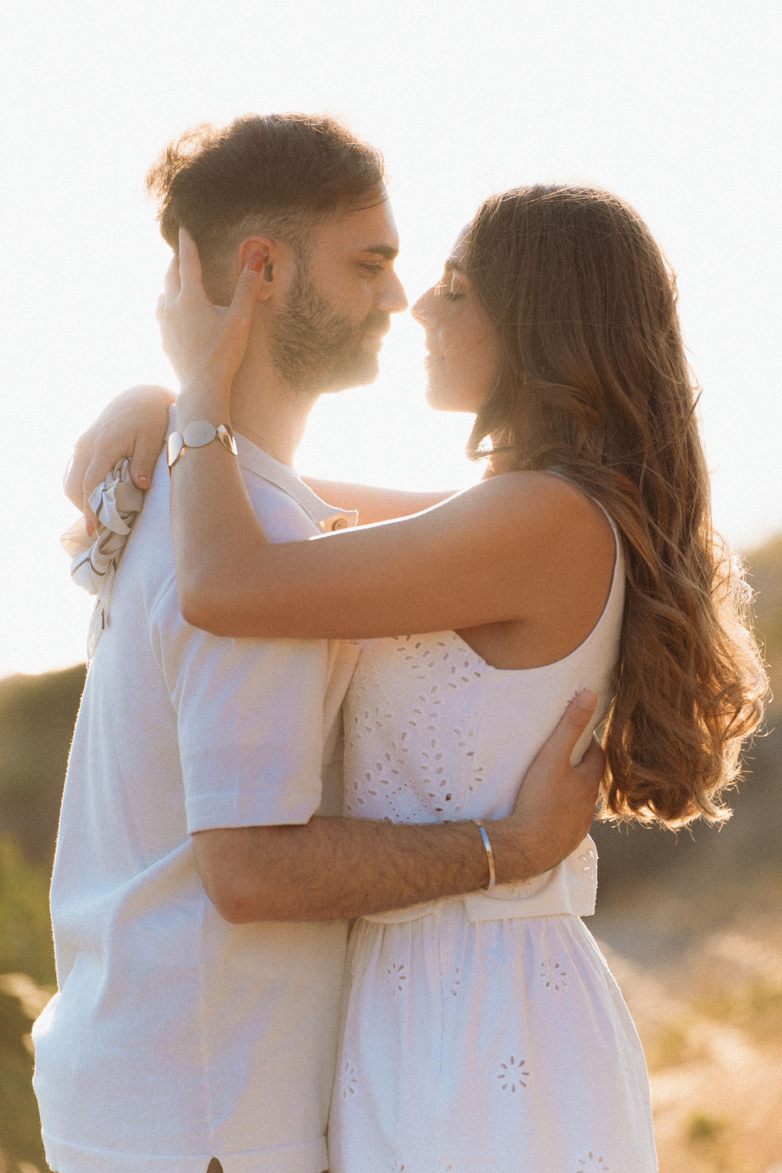 Young couple in white outfits sharing a tender moment, embracing face-to-face in warm golden sunlight outdoors.