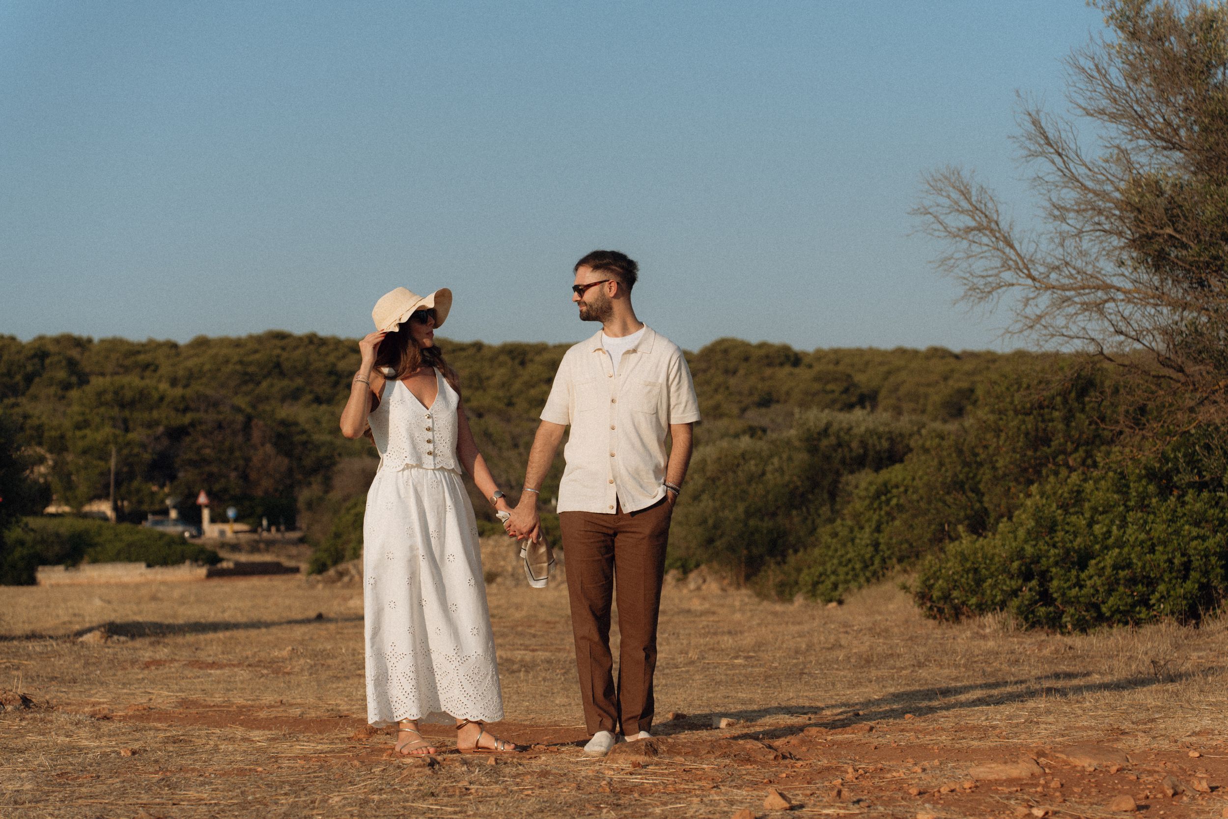 Couple holding hands in a sunny field, woman in white dress and hat, man in beige shirt and brown pants.