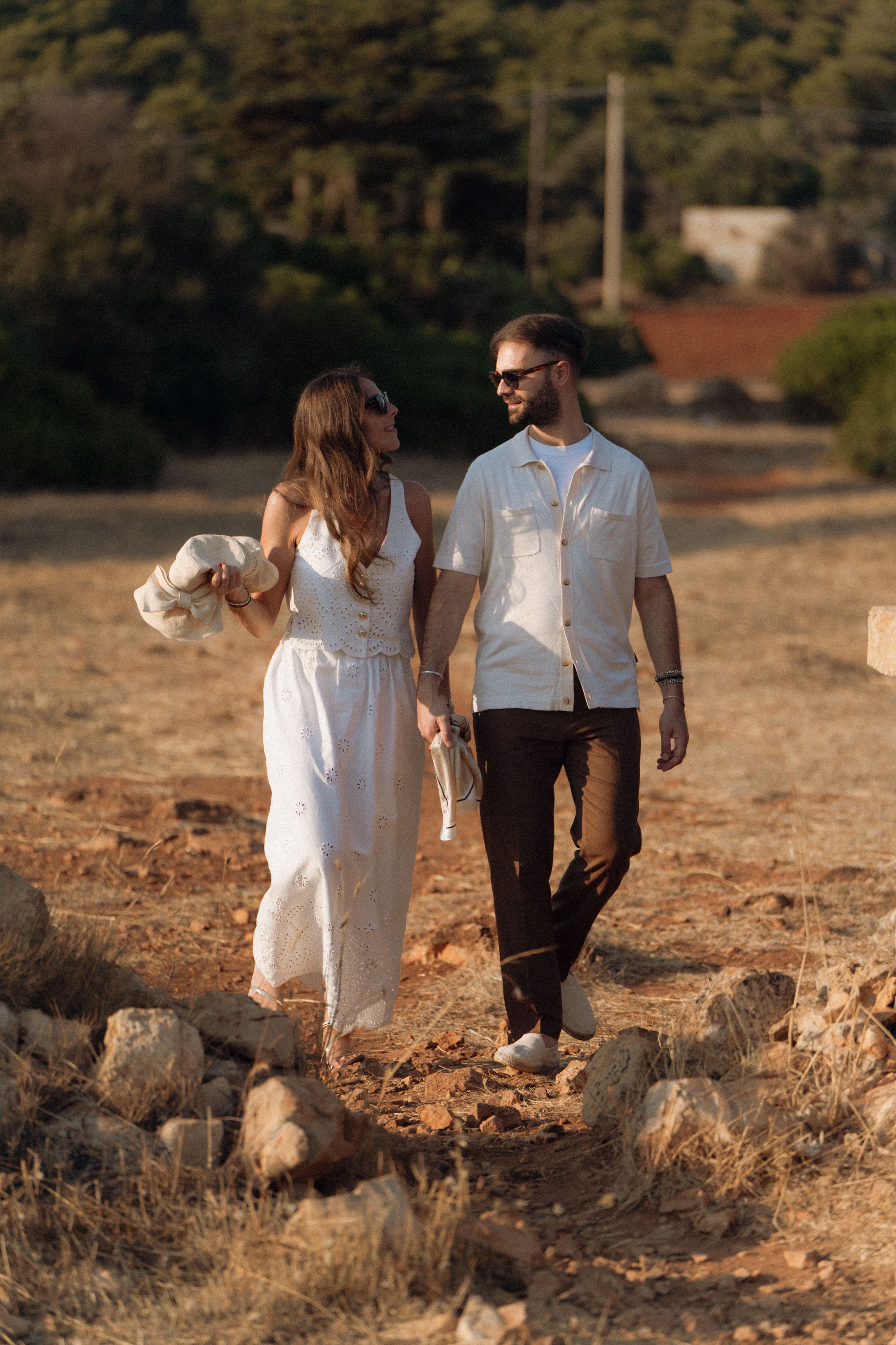 Couple walking hand in hand through a sun-drenched rocky field, woman in white dress, man in light shirt.