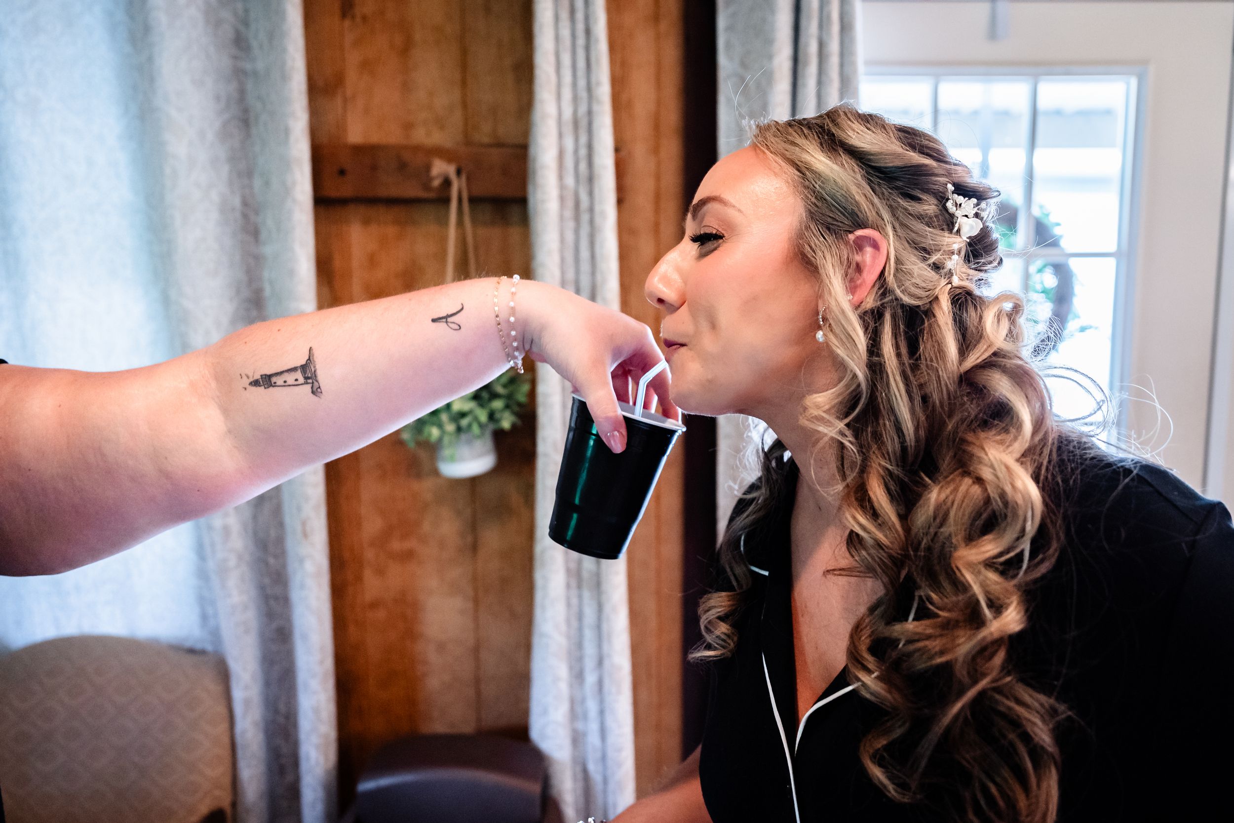 Bride sipping from a black cup during wedding preparations while someone with a tattoo holds it for her.