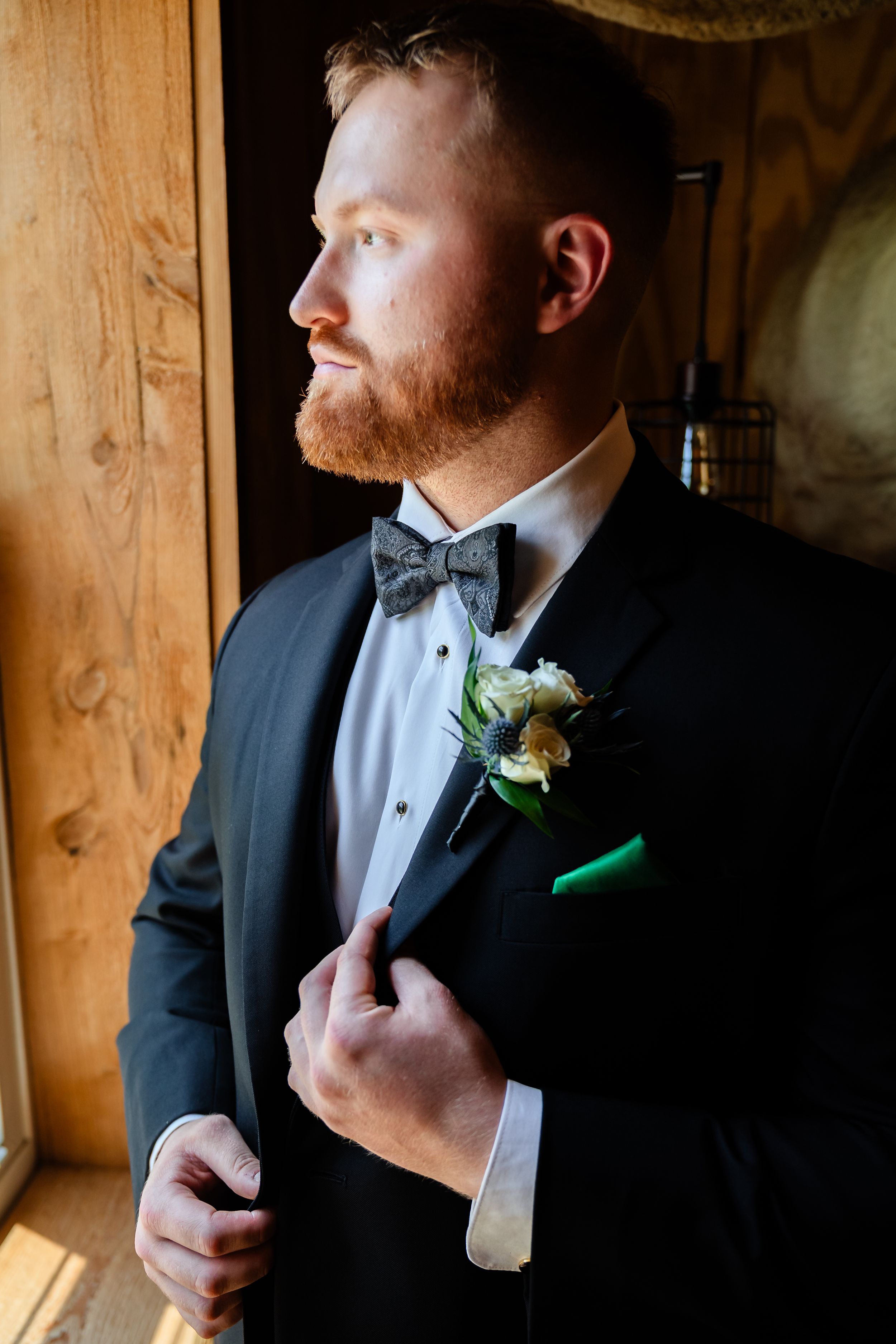 Dapper groom with red beard in black tuxedo and bow tie with boutonniere gazing out window on wedding day.