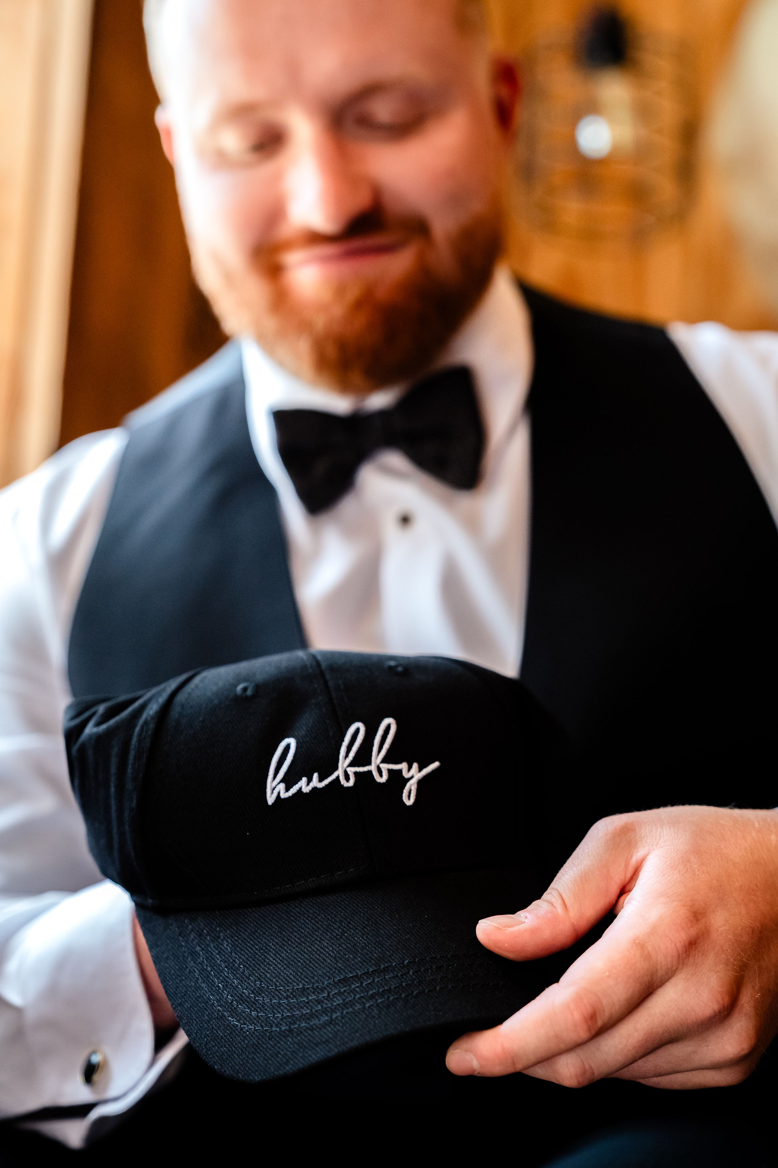 Groom in black tuxedo vest and bow tie holds a black cap embroidered with hubby in white script.