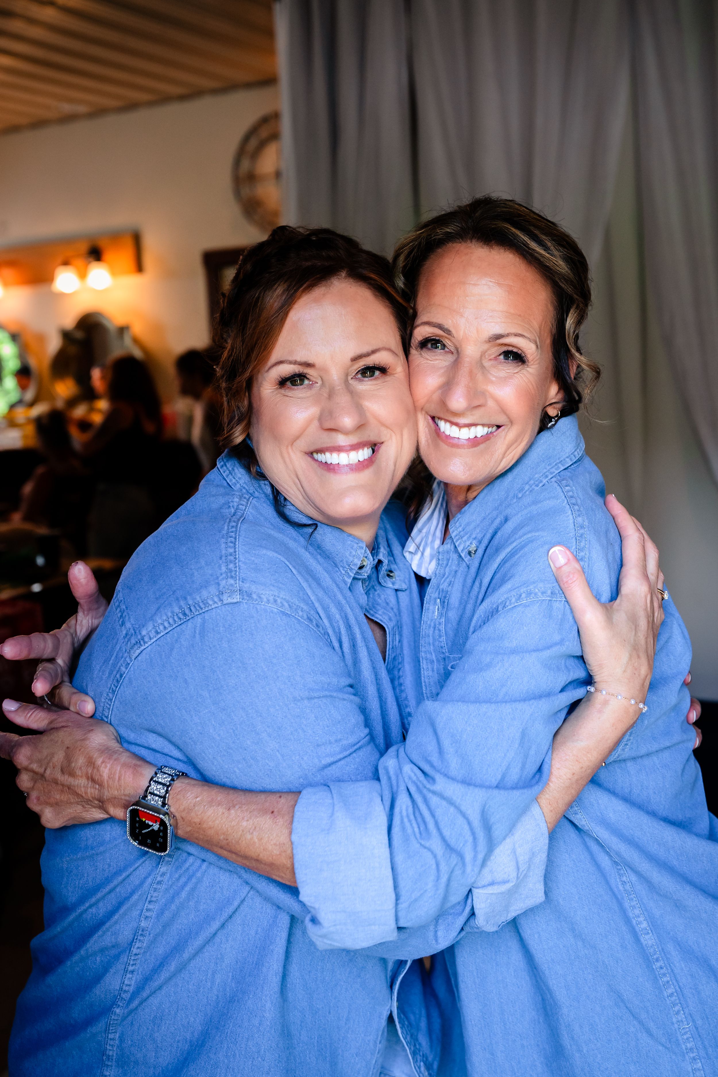 Two women in matching blue denim shirts share a warm hug and smile at a wedding celebration.