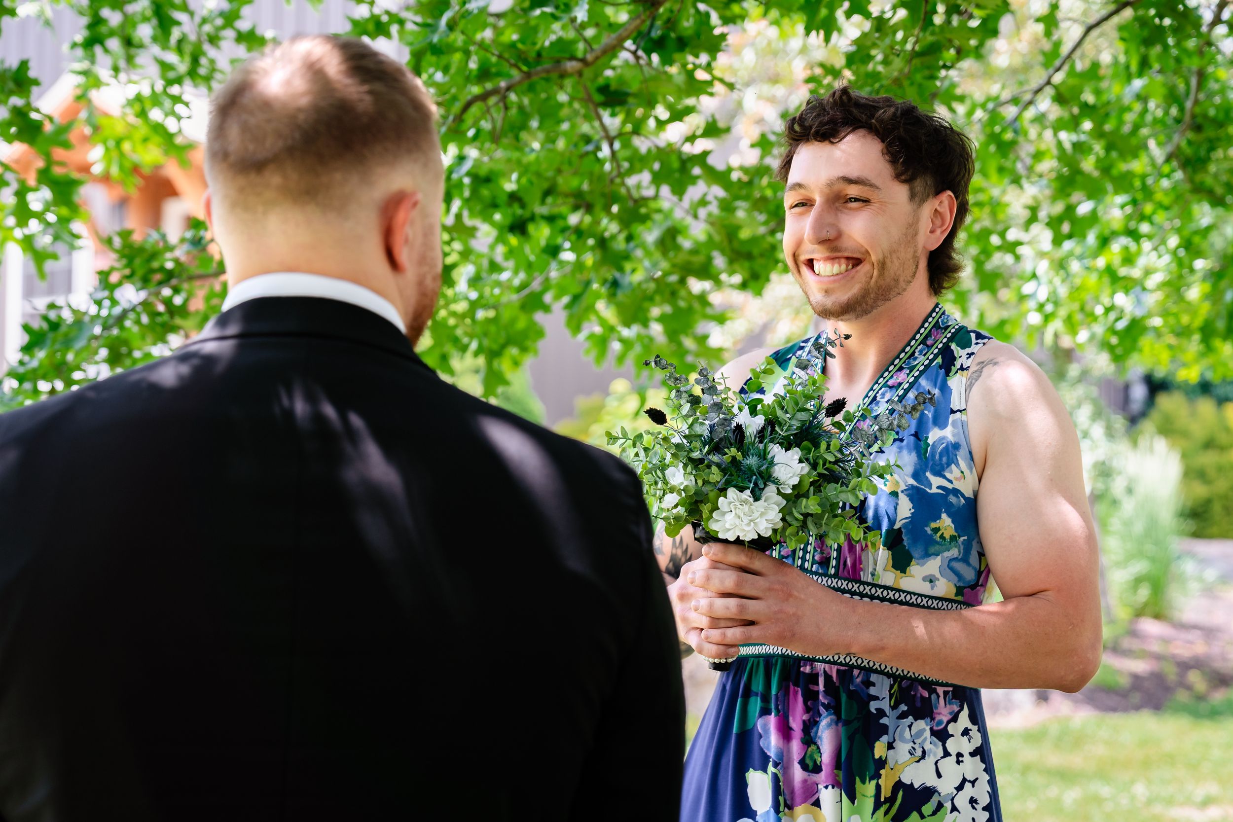 Smiling groomsman in floral dress holds a bouquet of white flowers while greeting a guest at an outdoor wedding.
