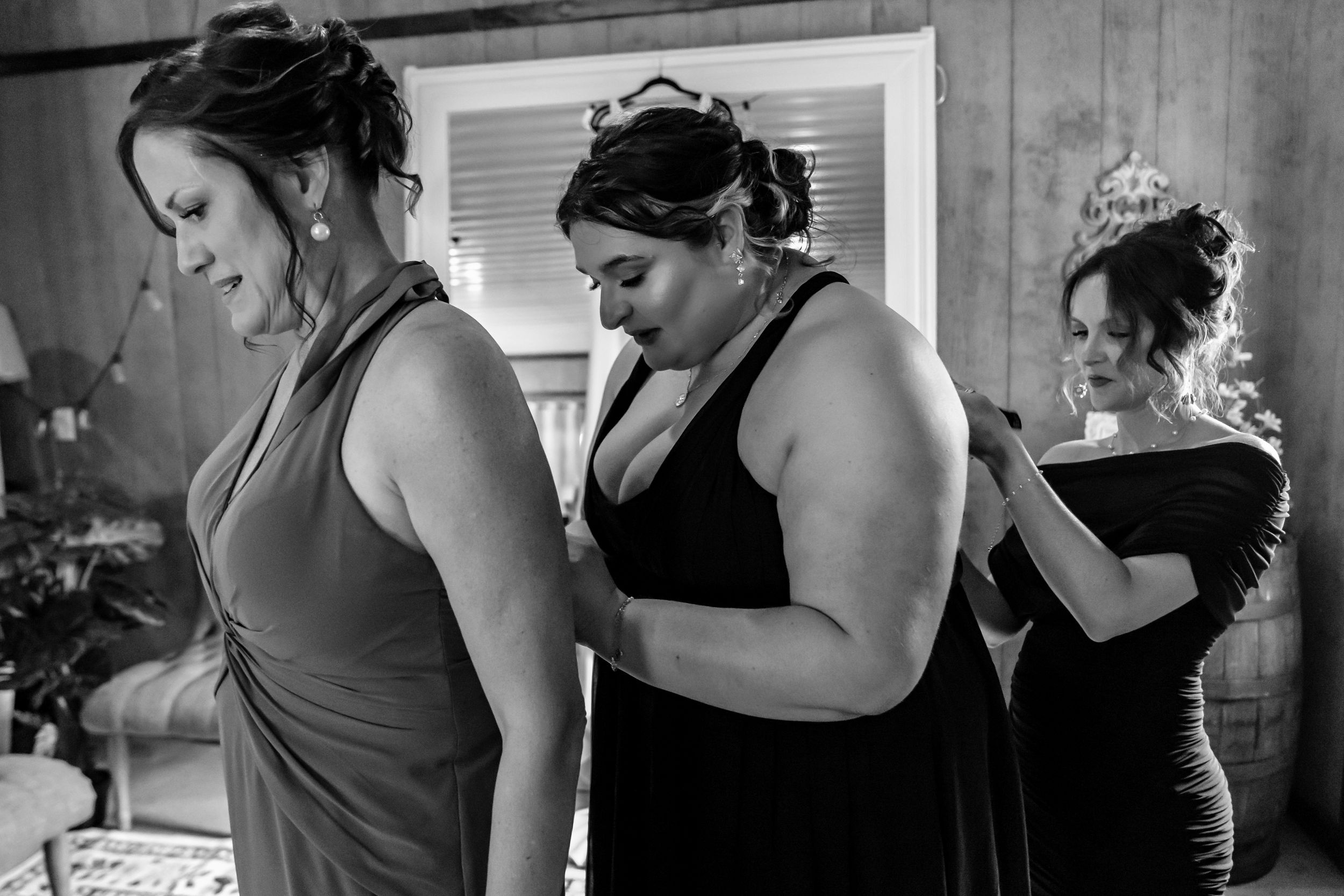 Three bridesmaids in black dresses help each other get ready in a rustic wooden room in black and white photo.