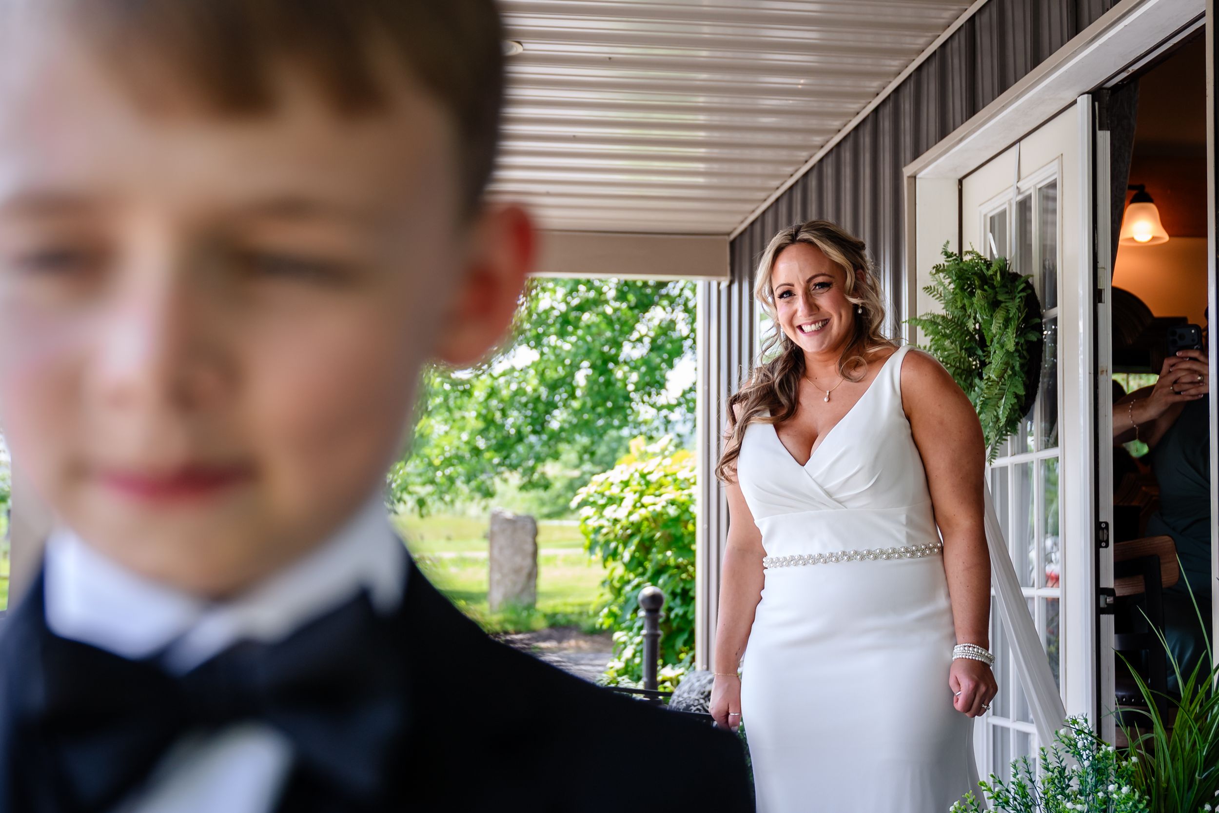 Smiling bride in white gown stands in doorway as young boy in tuxedo looks on during wedding day preparations.